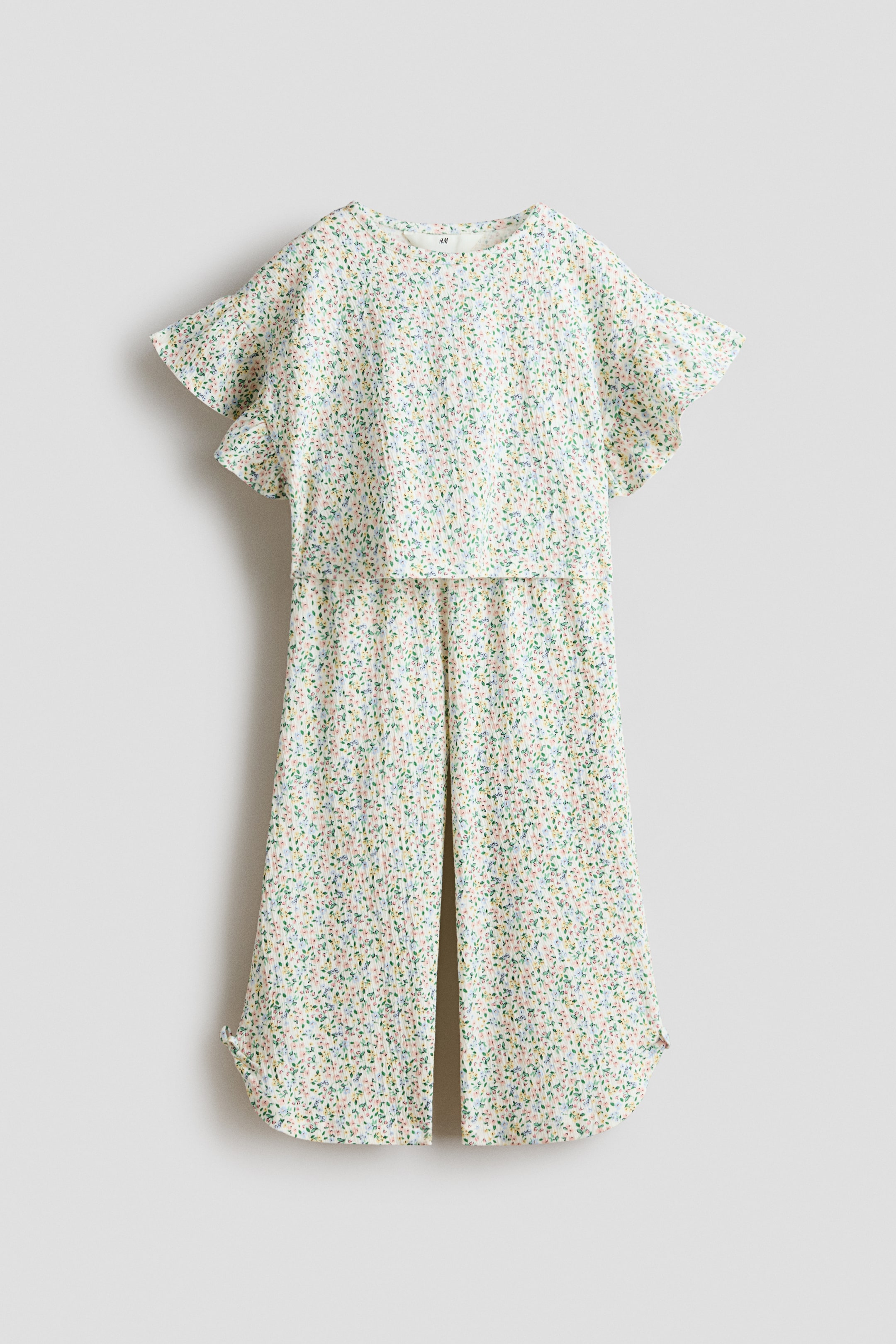 Ampliar la imagen: Un conjunto blanco de dos piezas, compuesto por un top y pantalones de pierna ancha, presenta un patrón moteado multicolor y una textura arrugada. El top cuenta con cuello redondo y mangas cortas con vuelo, mientras que los pantalones ofrecen un corte relajado con dobladillo ligeramente curvado, mostrados de frente (FRONT).