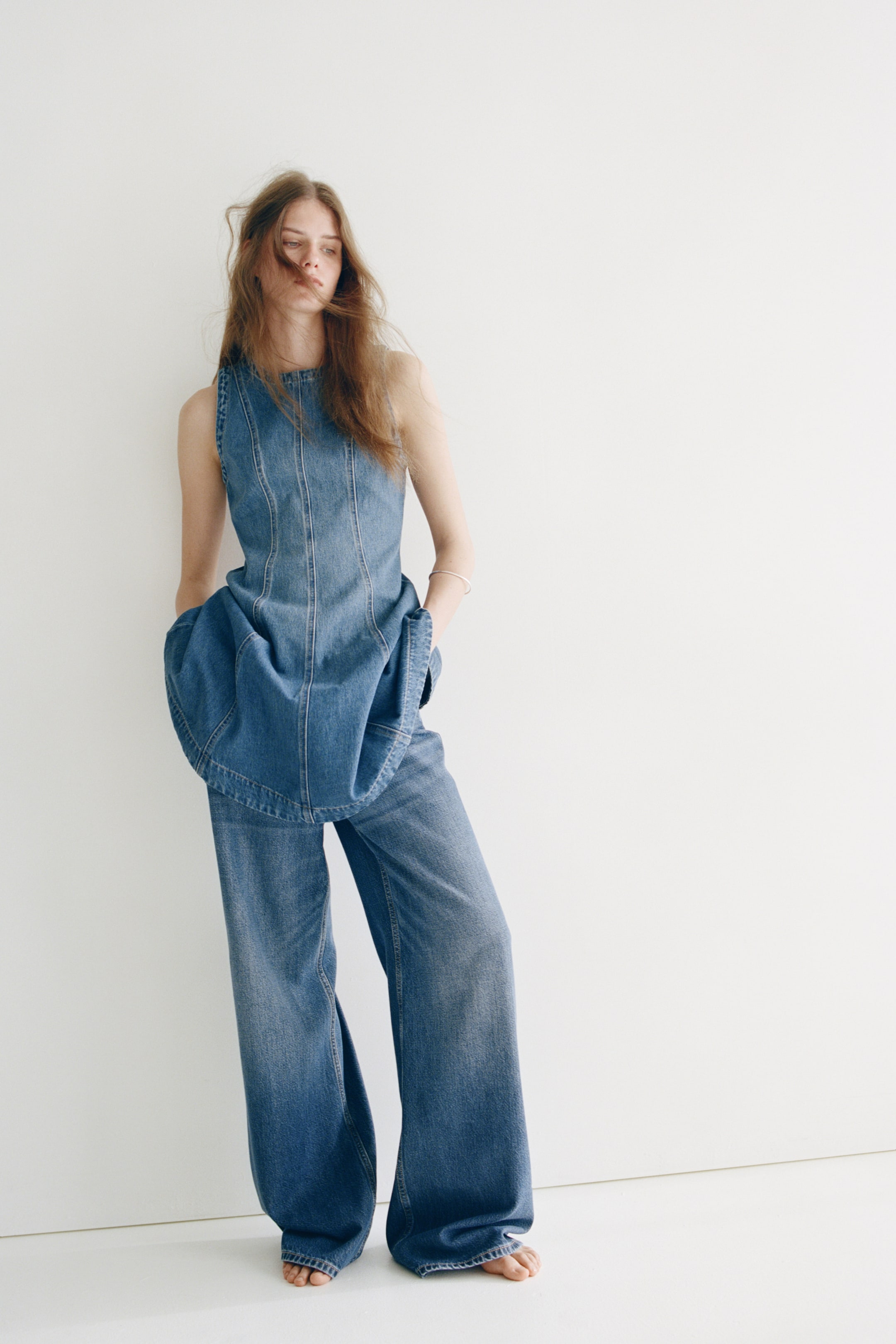 View larger image: A short, sleeveless dress in cotton denim, featuring a round neckline, fitted bodice, and a flared skirt with visible front seams, is being worn by the model. The dress is layered over wide-leg denim trousers, with the model's hands resting in the trousers' pockets.