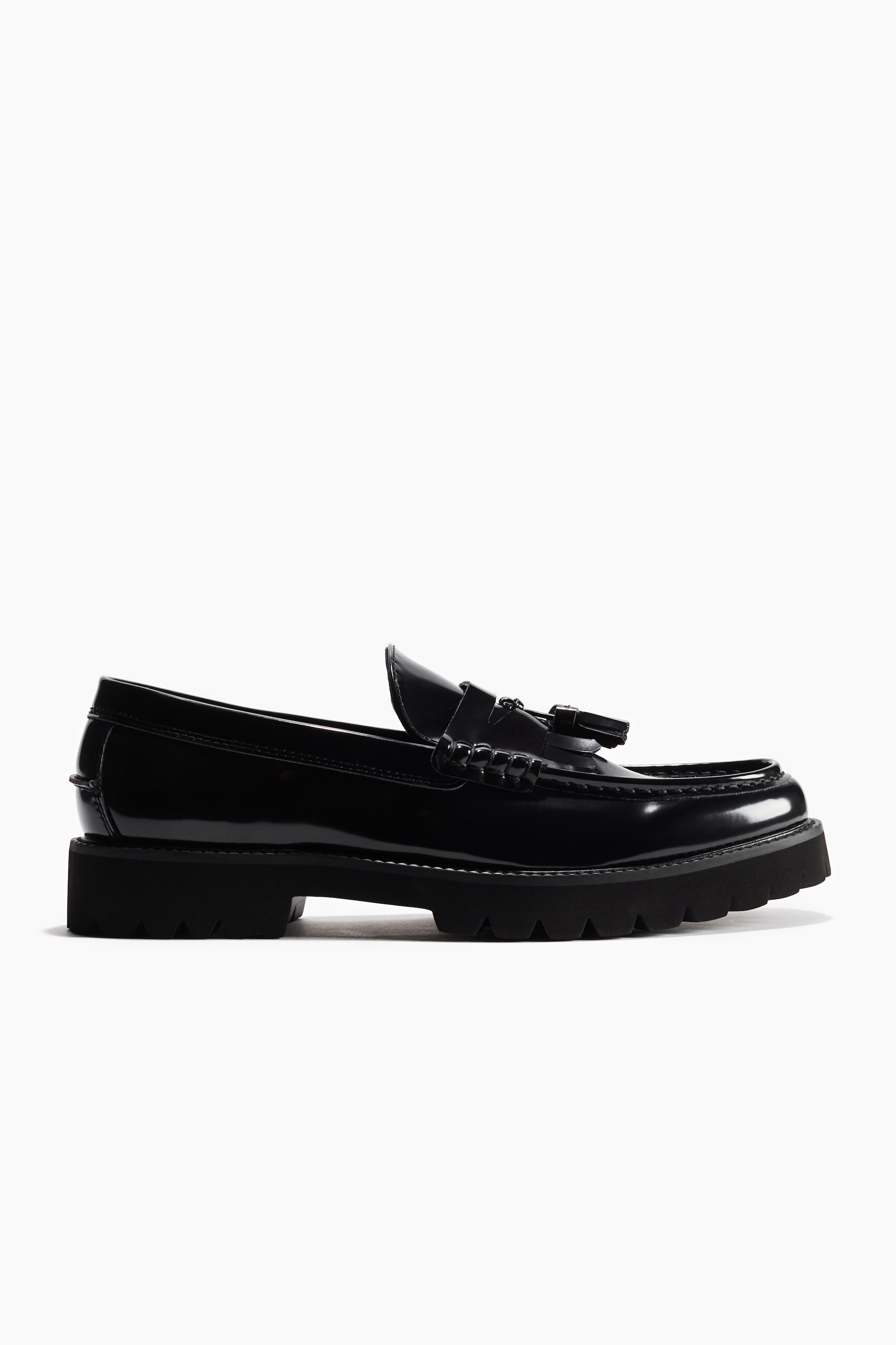 View larger image: A black patent leather tassel loafer is presented from a side angle, featuring a chunky lug sole and visible moc toe stitching on the vamp.