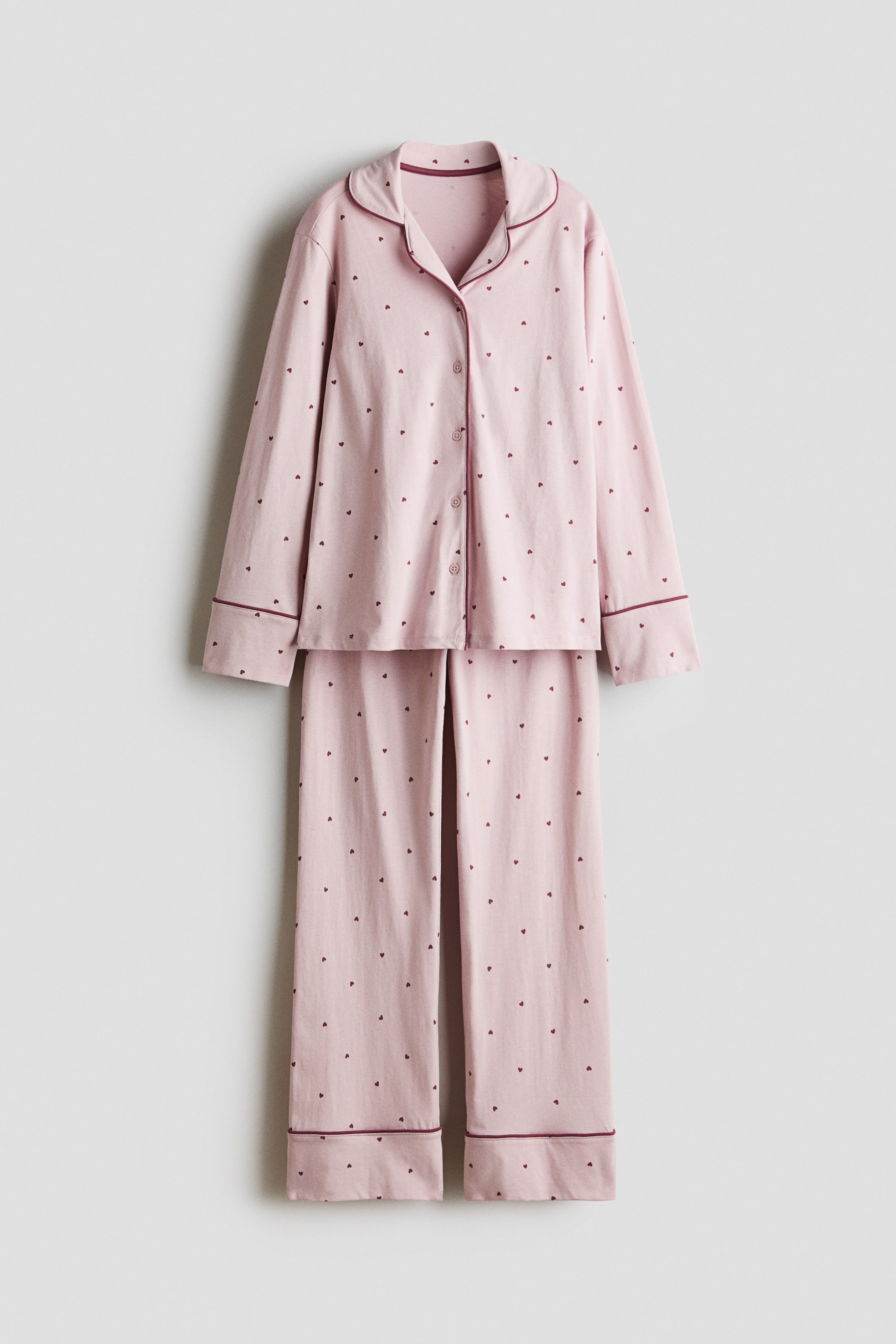 View larger image: A front-facing view showing a light pink cotton jersey pyjama set with a small dark red heart pattern, consisting of a long-sleeved shirt and matching long bottoms. The shirt features a resort collar, front button closure, and cuffed sleeves, with both pieces displaying contrasting dark red piping along the collar, placket, and cuffs.