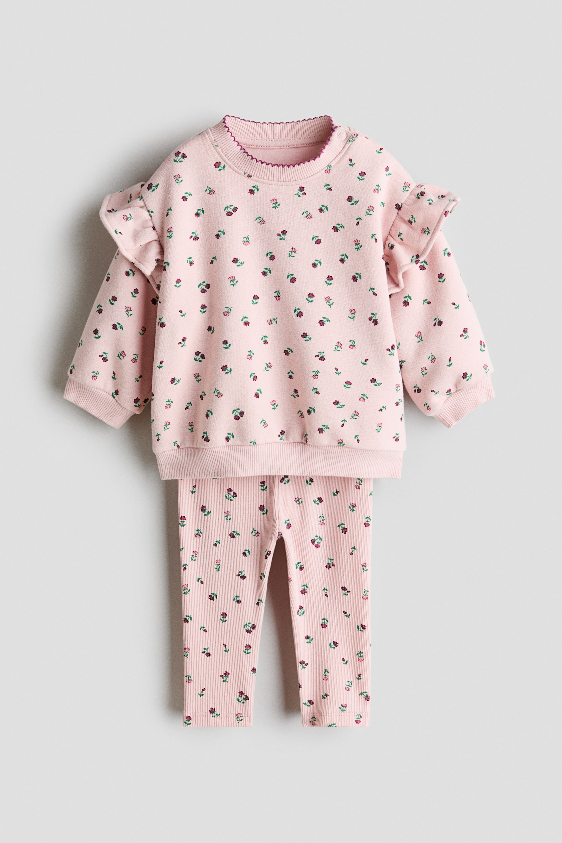 View larger image: A pink two-piece baby set, shown from the front, featuring a sweatshirt and matching ribbed leggings. The sweatshirt has a small floral print, ruffled shoulder details, ribbed cuffs and hem, and a scalloped trim along the neckline, while the leggings also feature the floral print.