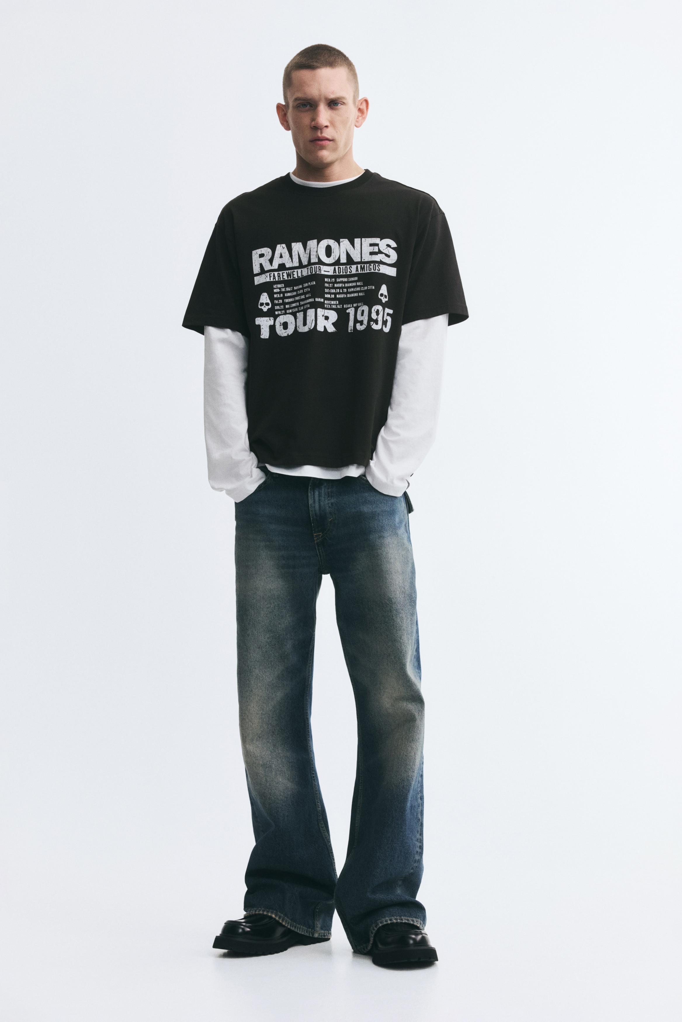 A black midweight cotton jersey t-shirt is showcased from the front, featuring a distressed white graphic print on its front that displays 'RAMONES FAREWELL TOUR - ADIOS AMIGOS TOUR 1995,' along with tour dates and skull icons. It is designed with dropped shoulders and a round neckline with a narrow trim, offering a loose fit.