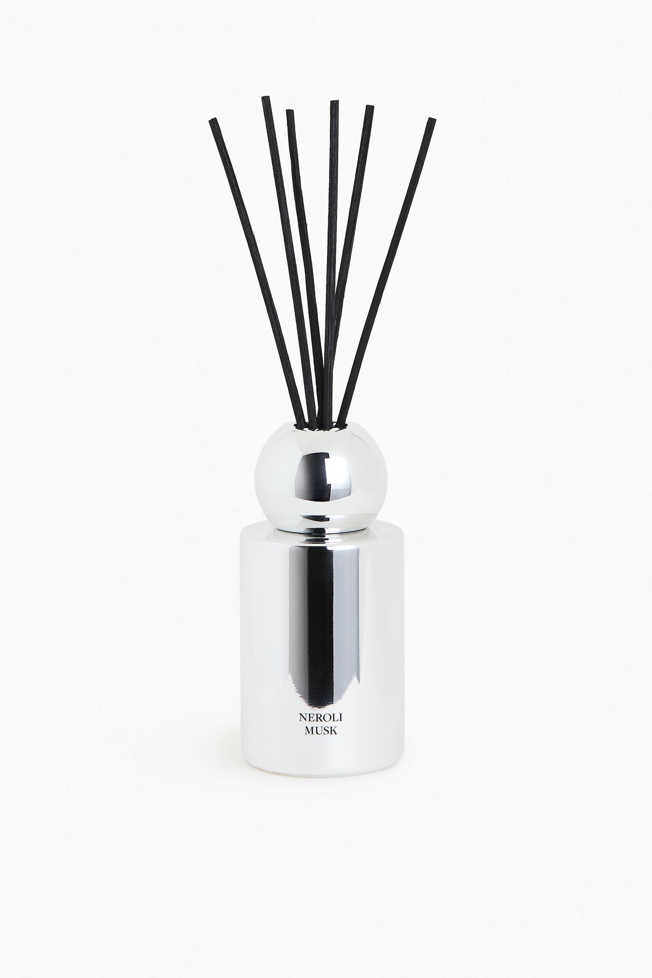 View larger image: A reed diffuser features a cylindrical silver container with a highly reflective, glossy finish, displaying the text 'NEROLI MUSK' on its front. A spherical silver cap tops the container, from which several black diffuser reeds emerge and fan upwards.