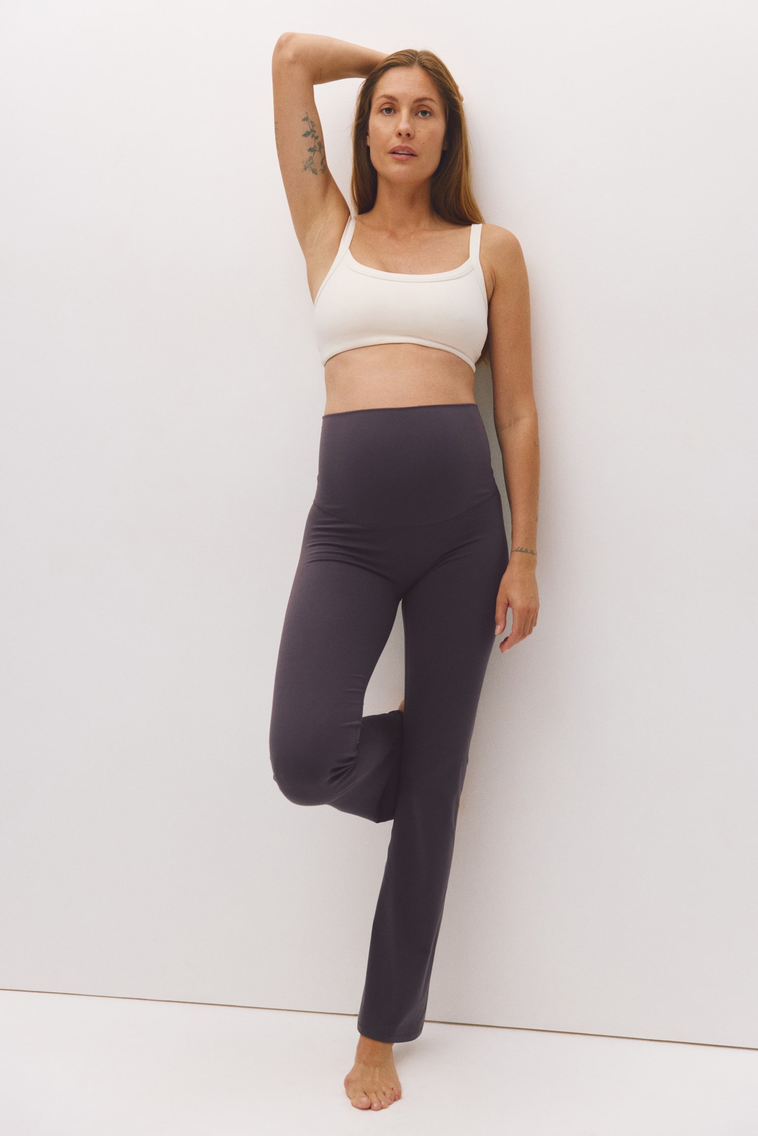 Dark grey maternity flare trousers featuring a high, over-the-bump waistband are being worn by the model. The trousers are styled with a white scoop-neck cropped top. - 1