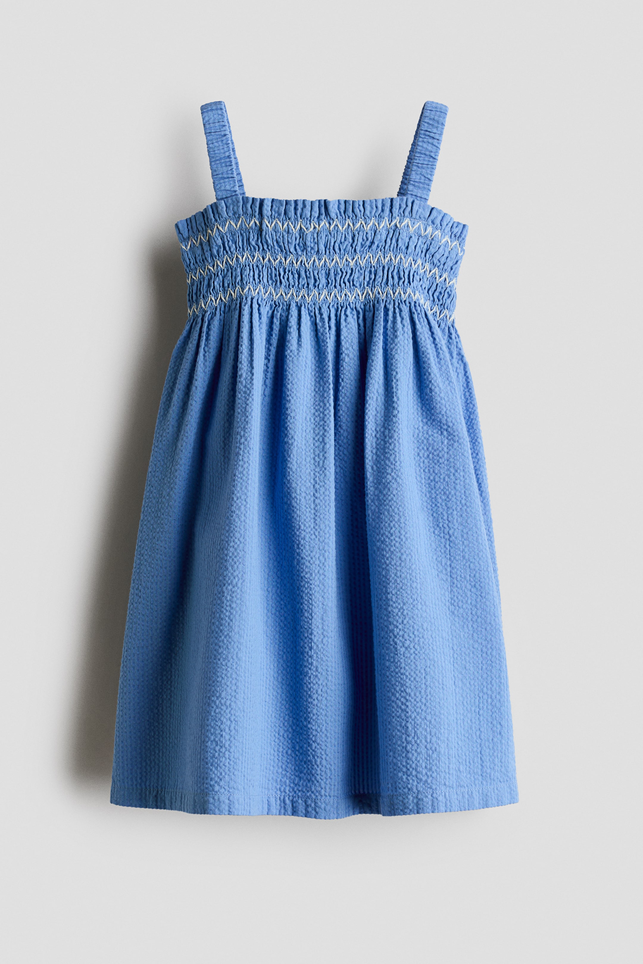 View larger image: A blue seersucker dress is hanging, facing the front, featuring thick shoulder straps, a smocked bodice with white decorative embroidery in a zigzag pattern, and a gathered, flared skirt.