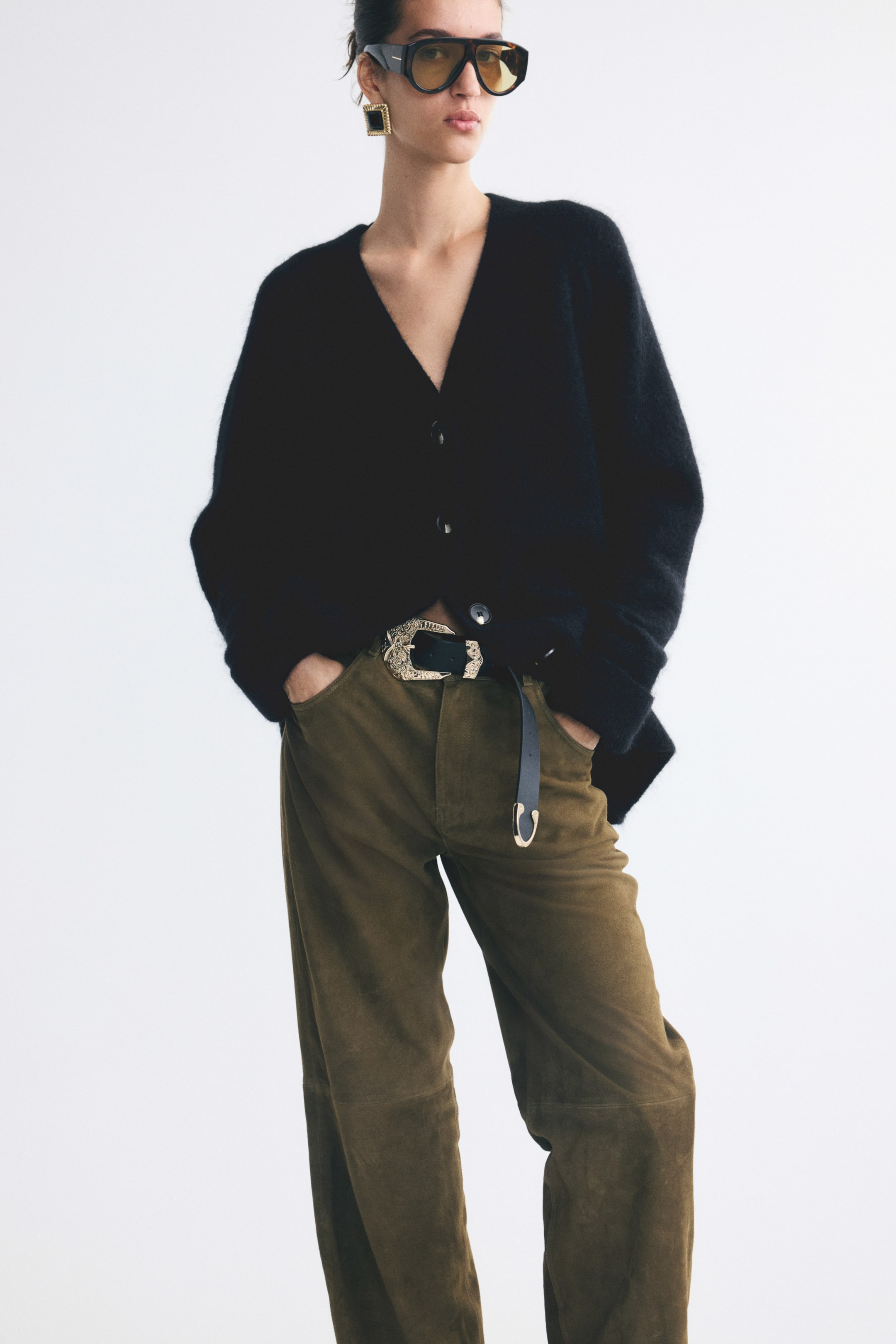 Größeres Bild anzeigen: Ein oversized schwarzer Cardigan in einem weichen, flauschigen Strick wird vom Model getragen. Der Cardigan verfügt über einen V-Ausschnitt, eine durchgehende Knopfleiste und lange Ärmel und ist am Saum teilweise offen getragen und mit braunen Hosen kombiniert.