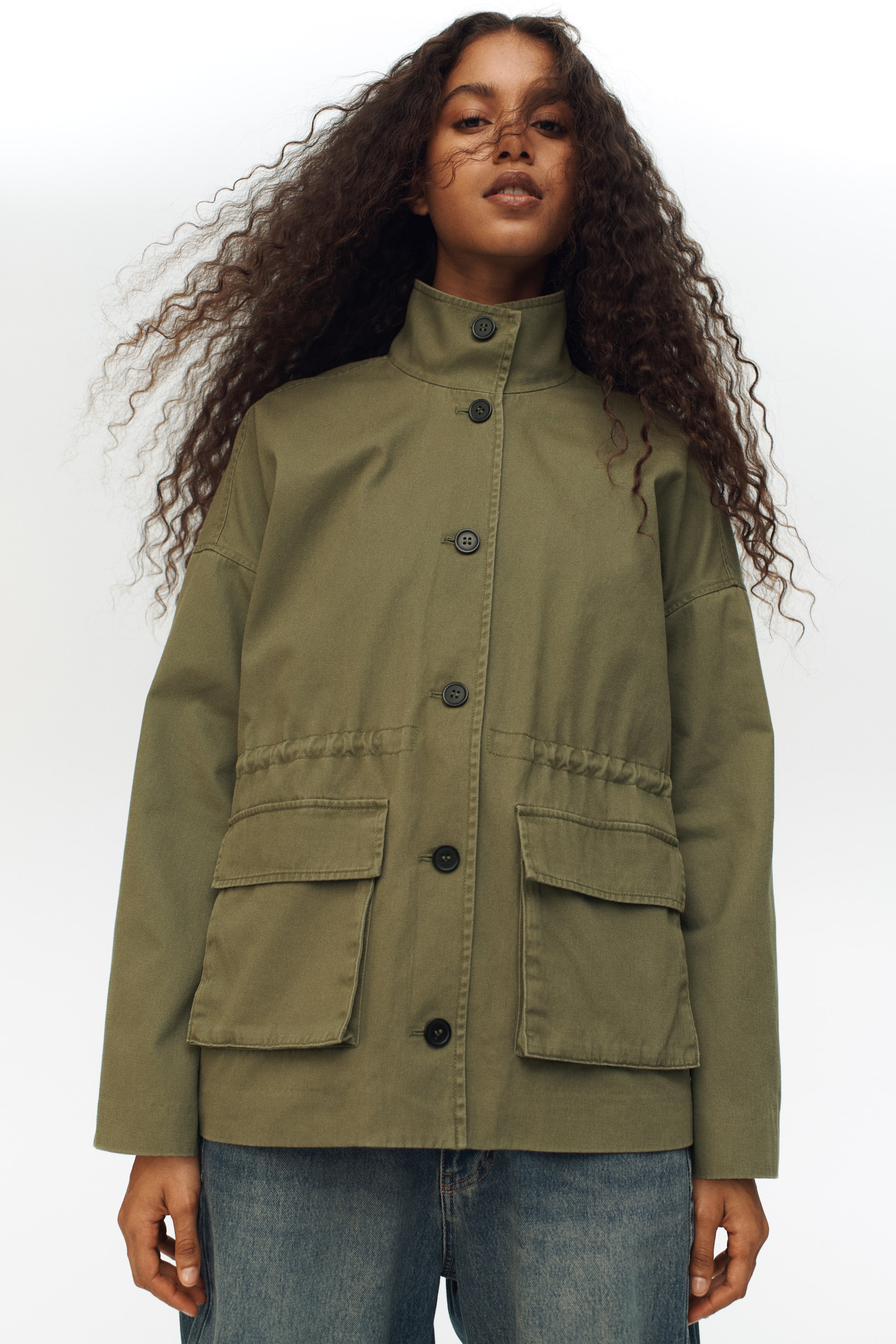 View larger image: The model is wearing an olive green cotton twill jacket, showcasing a funnel collar, buttoned front, and dropped shoulders. The jacket features bellows front pockets with flaps, and an internal drawstring that cinches the waist.