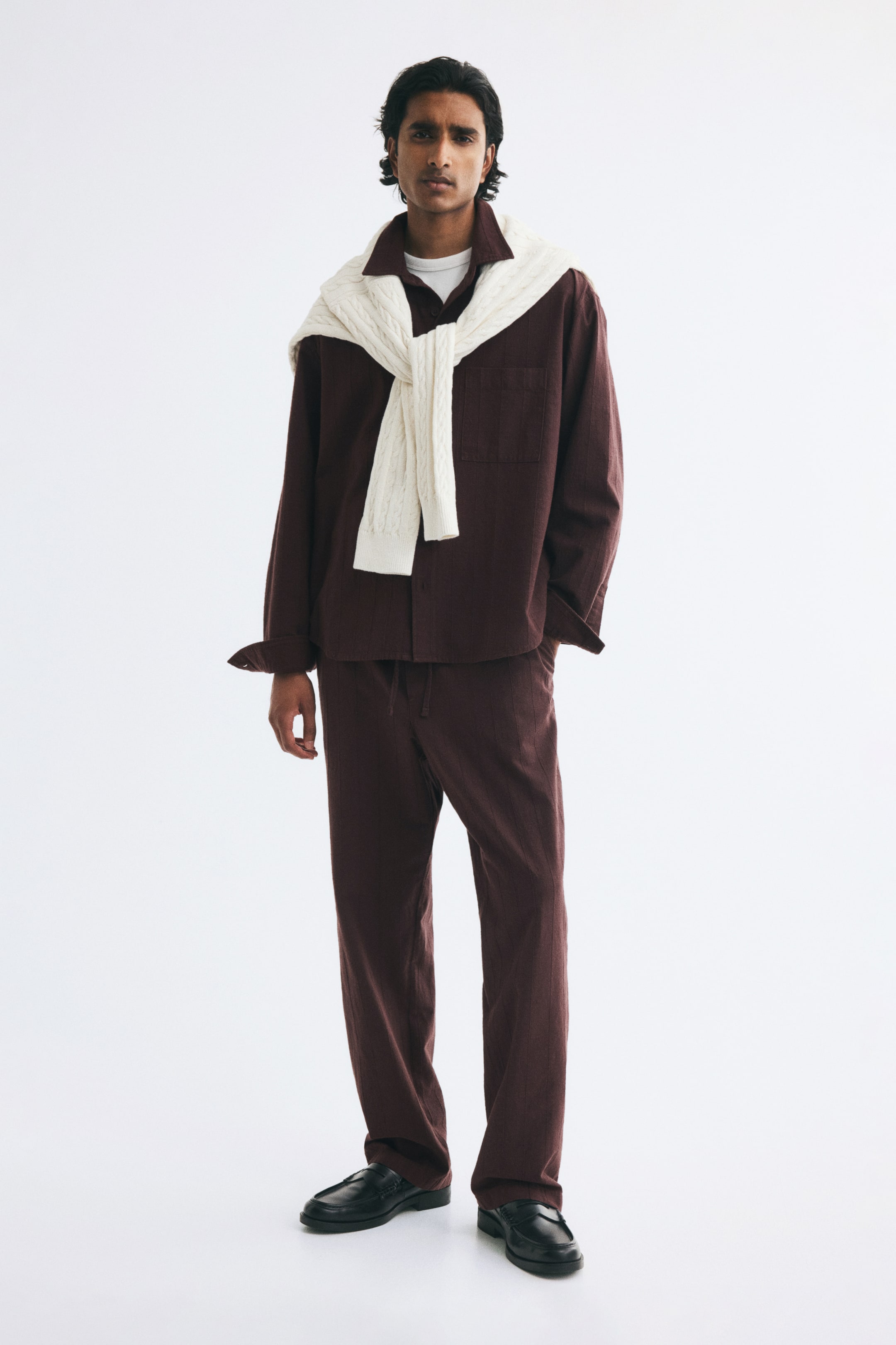 View larger image: A dark brown, relaxed-fit shirt in a cotton dobby weave is being worn by the model, styled with a cream-coloured cable-knit sweater draped over the shoulders and matching brown trousers. The shirt features a collar, a French front, an open chest pocket, and long sleeves with cuffs.