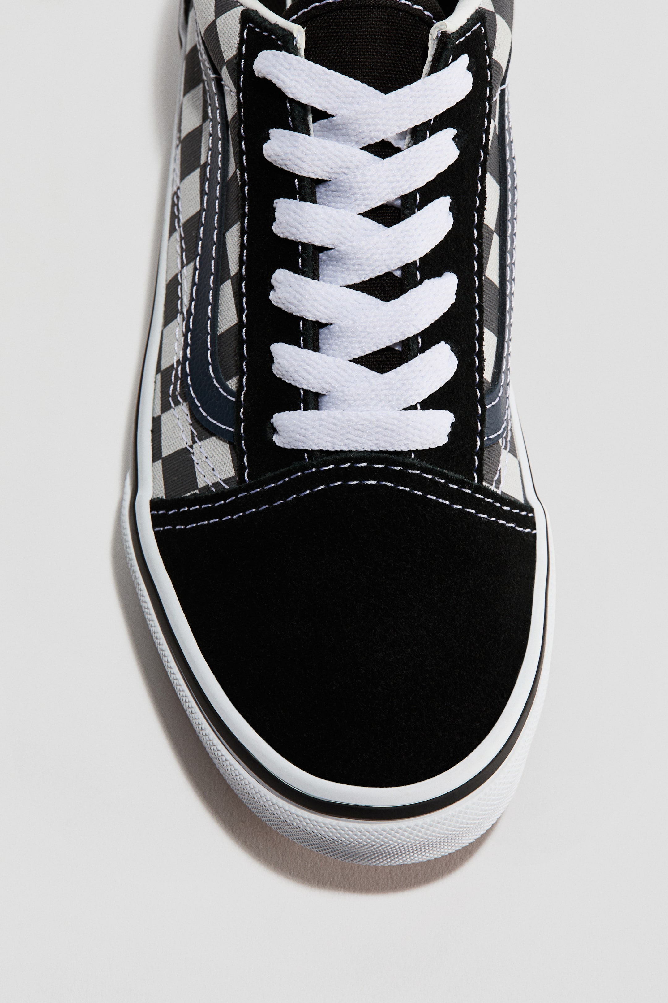 Größeres Bild anzeigen: Ein Sneaker präsentiert seine Vorderseite, mit einer schwarzen Wildleder-Zehenkappe und weißen, sich kreuzenden Schnürsenkeln. Der Mittelfuß zeigt ein schwarzes und graues Schachbrettmuster aus Canvas mit einem dunklen Seitenstreifen, der auf einer weißen Gummisohle mit einer schwarzen Linie ruht.