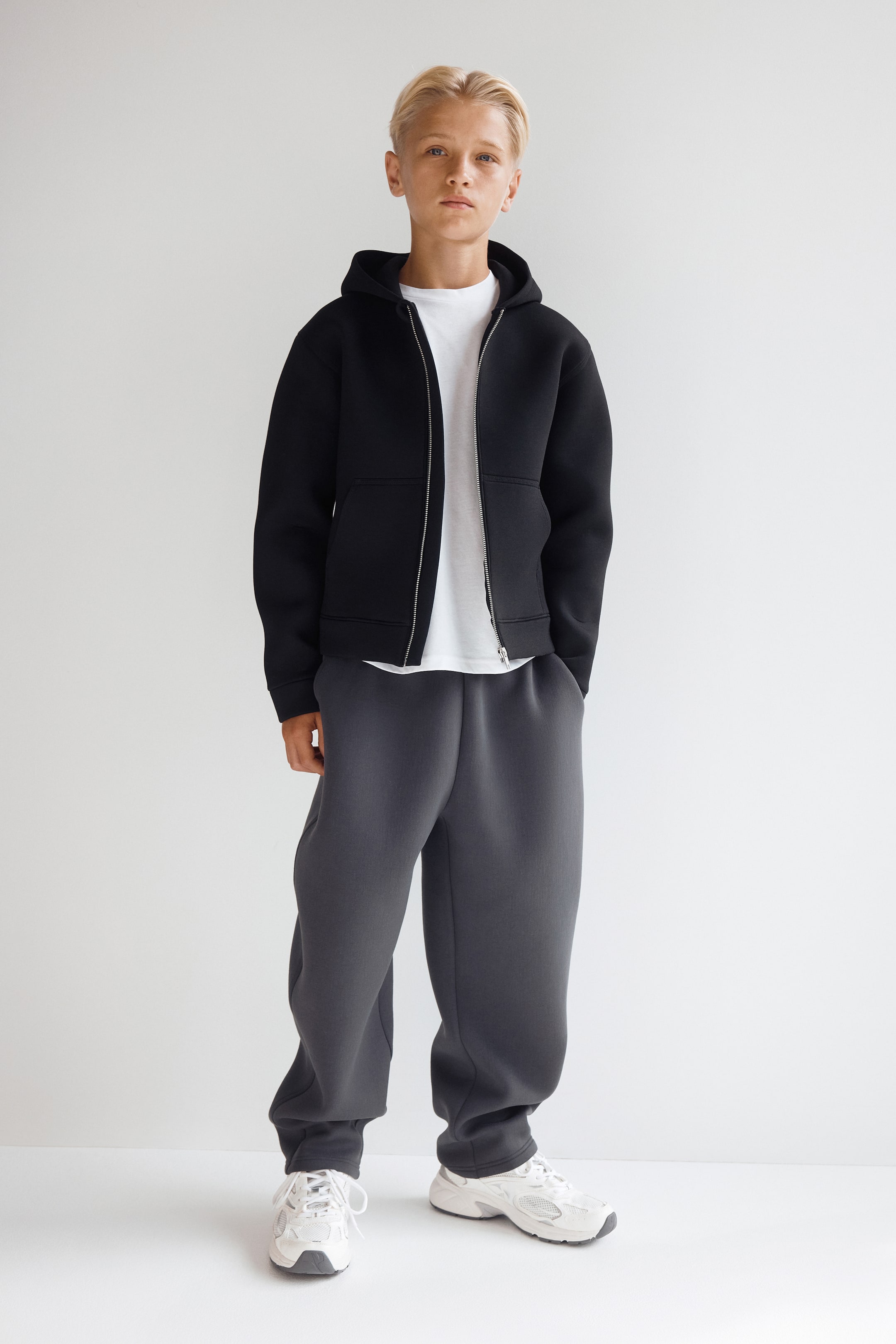 View larger image: A black zip-up hoodie and dark grey sweatpants are shown from the front, showcasing a relaxed, casual fit. The hoodie features a hood, a silver front zip fastening and open split kangaroo pockets, worn over a white T-shirt, while the sweatpants feature a loose silhouette with gathered ankle cuffs.