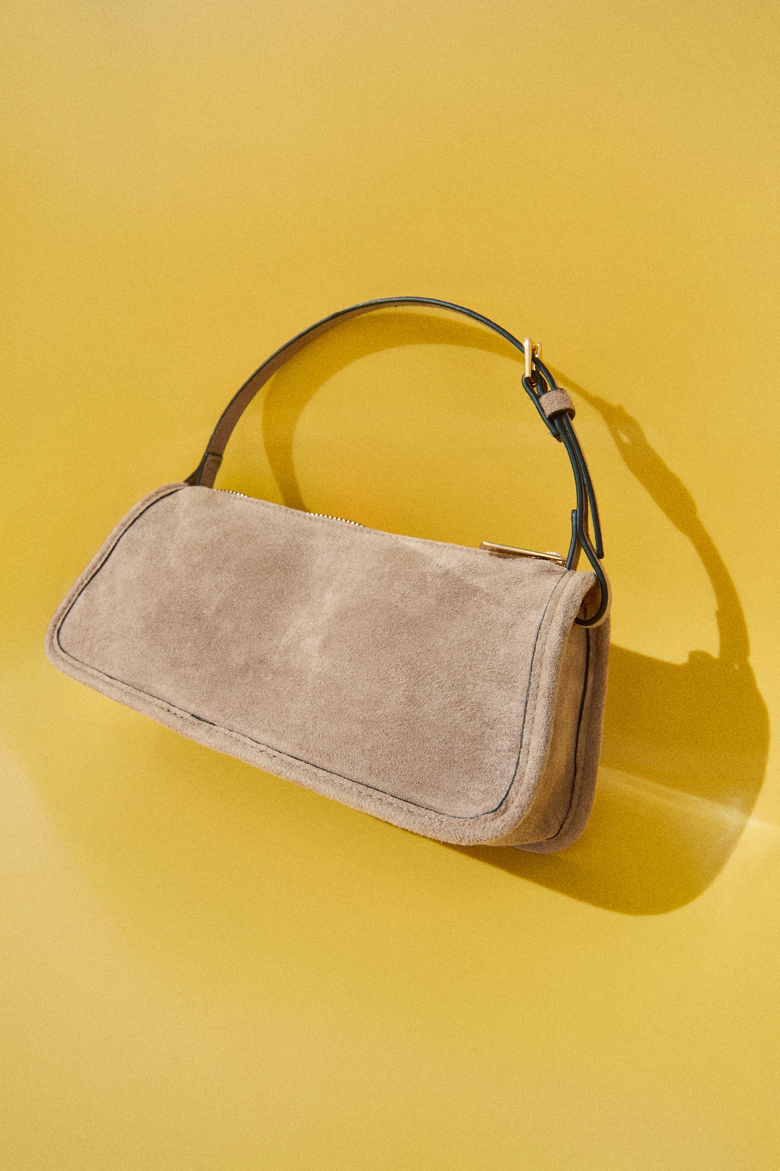A light brown suede handbag is showcased at the front, featuring a gold-toned top zip and a slender black adjustable strap. - 1