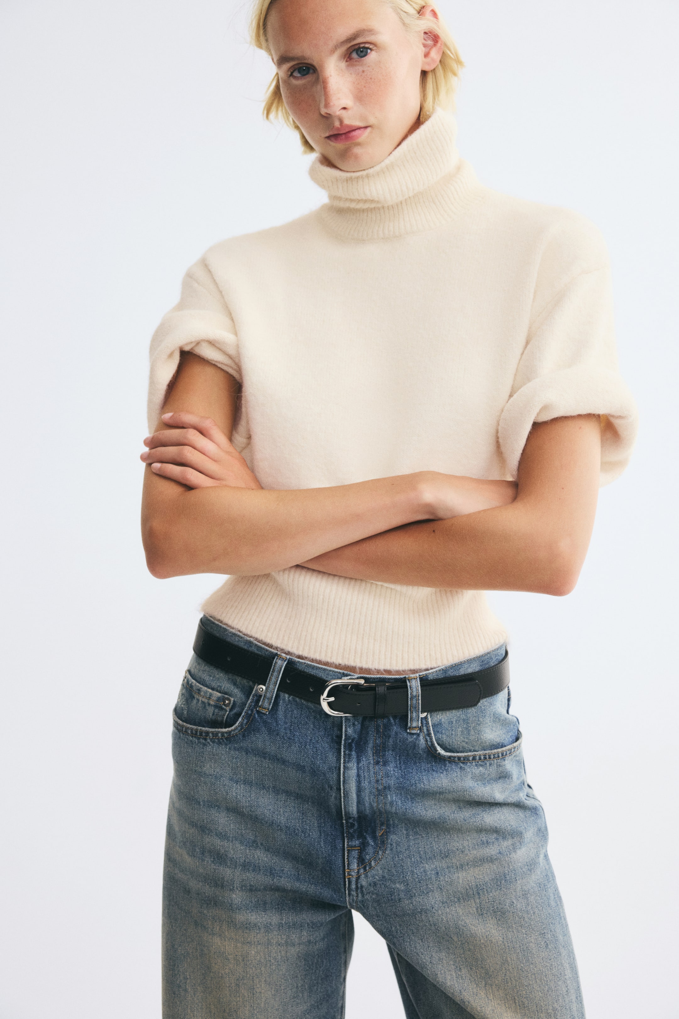 Agrandir l'image: Un pull à manches courtes en maille douce de couleur crème, doté d’un col montant côtelé, de épaules tombantes et d’un ourlet côtelé, est porté par le modèle. Le modèle se tient debout, les bras croisés, et associe le pull à un jean bleu et une ceinture noire.