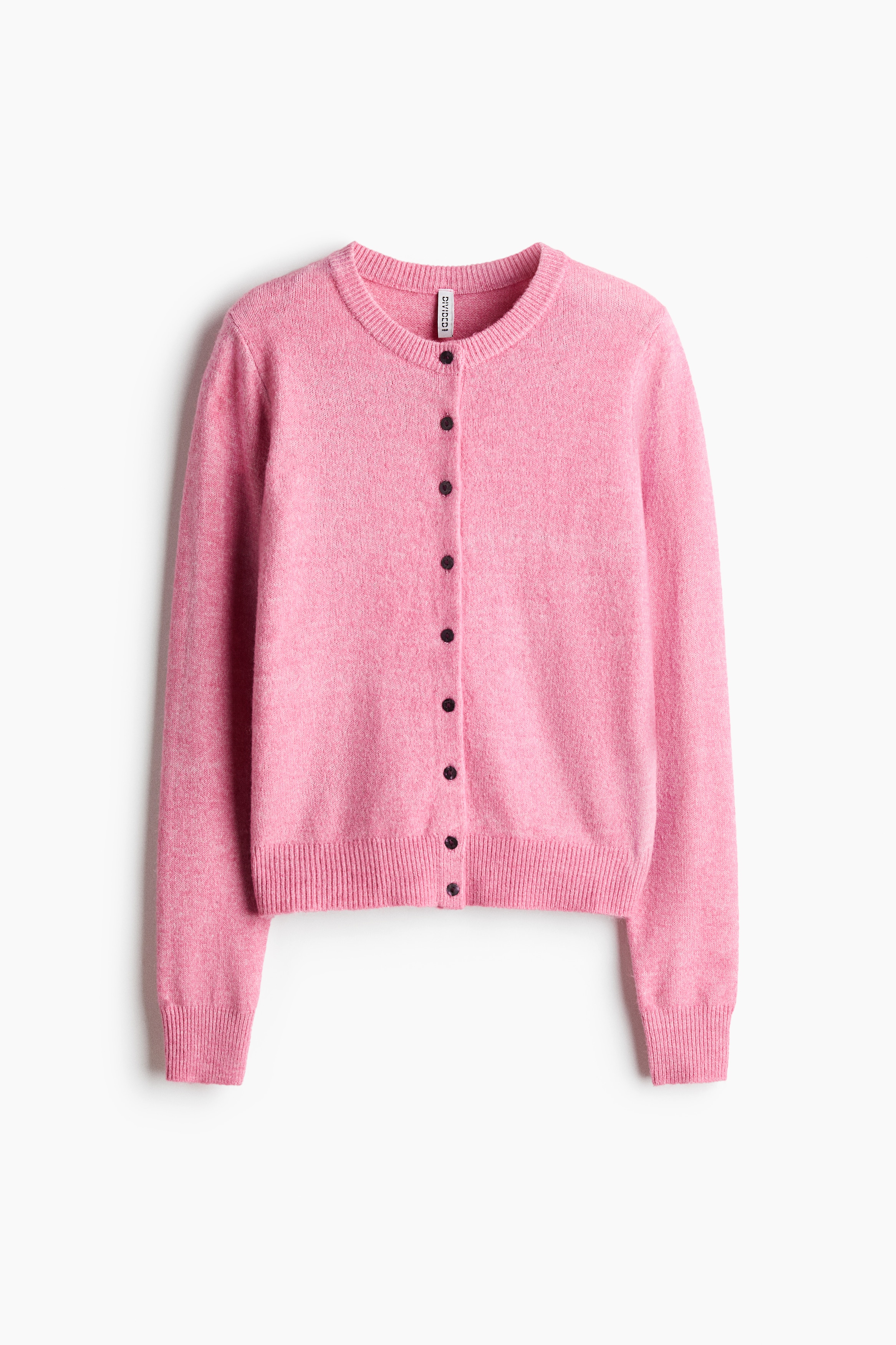A pink, fitted cardigan in a soft knit is being worn by the model, featuring a round, rib-trimmed neckline, dark front buttons, and ribbing at the cuffs and hem. The cardigan is styled partially unbuttoned at the bottom with blue jeans and a black belt, with the model standing with hands in the pockets.