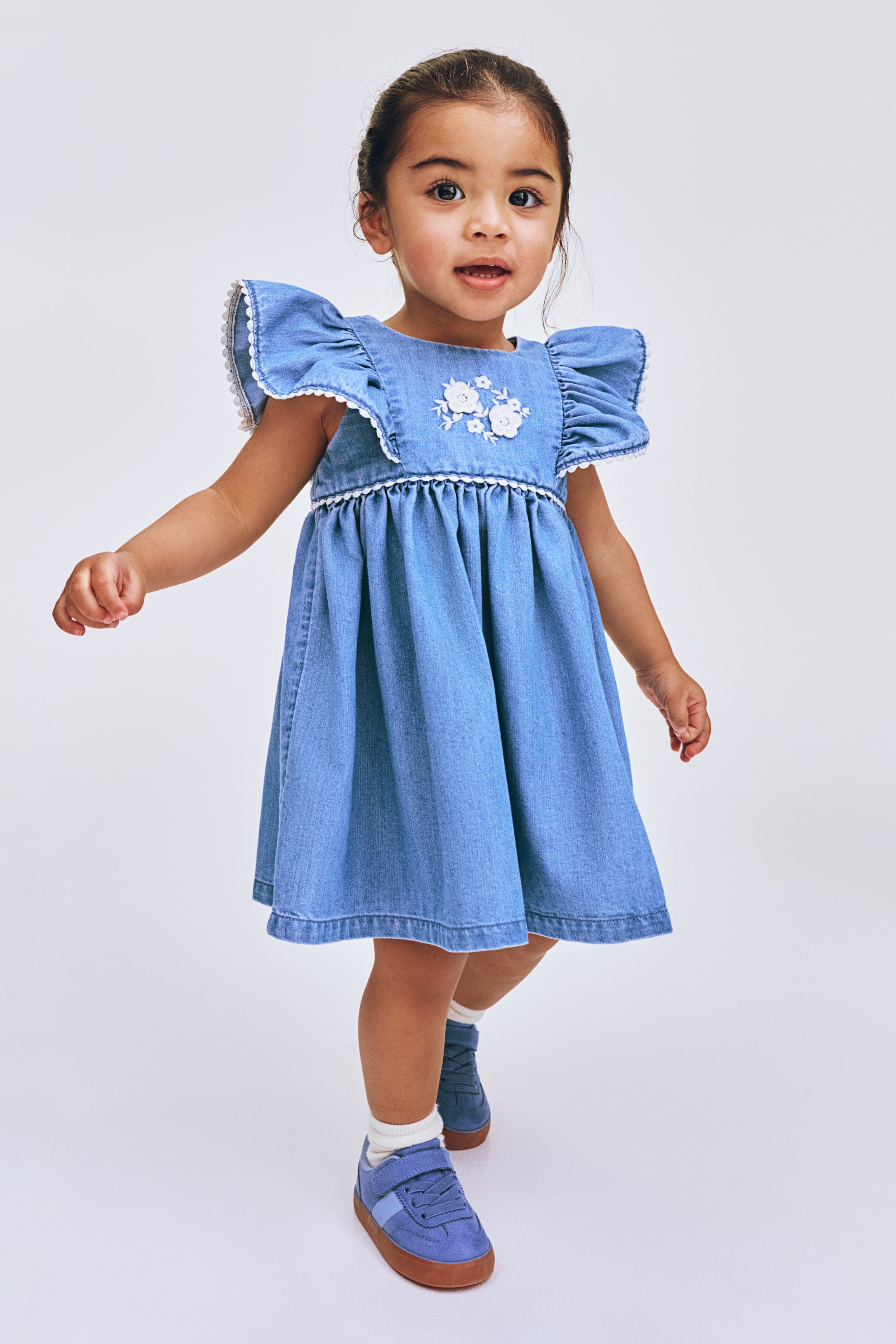 View larger image: This light blue denim dress showcases ruffled cap sleeves adorned with white lace trim. The chest is elegantly detailed with white floral embroidery, while a white trim accentuates the gathered waistline of the A-line skirt.