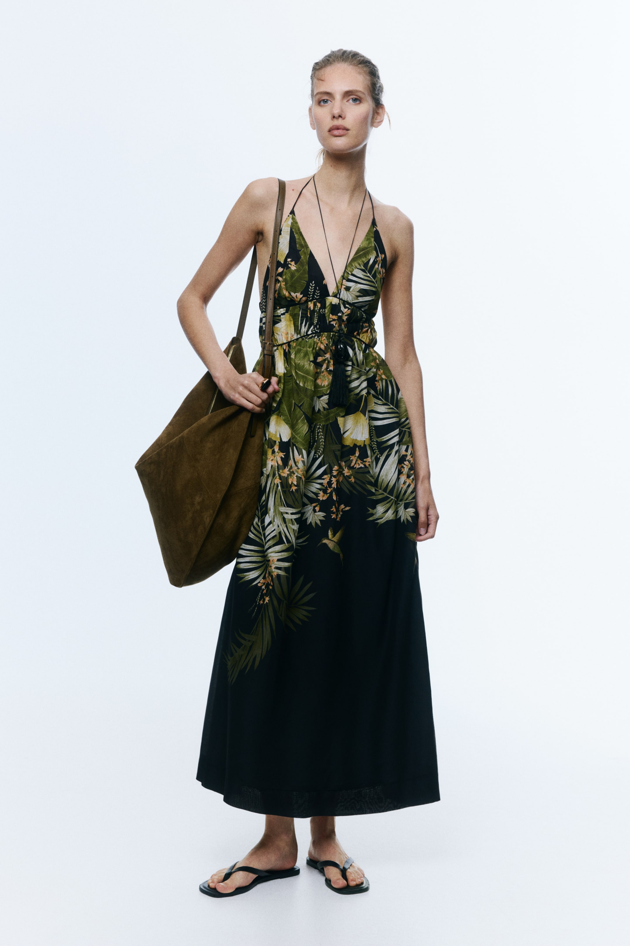 Agrandir l'image: La robe longue noire présente un col licou, un décolleté en V profond et une taille cintrée ornée de pompons noirs. Elle affiche un imprimé tropical de feuilles vertes et jaunes et de colibris, s'étendant jusqu'à une longueur maxi.