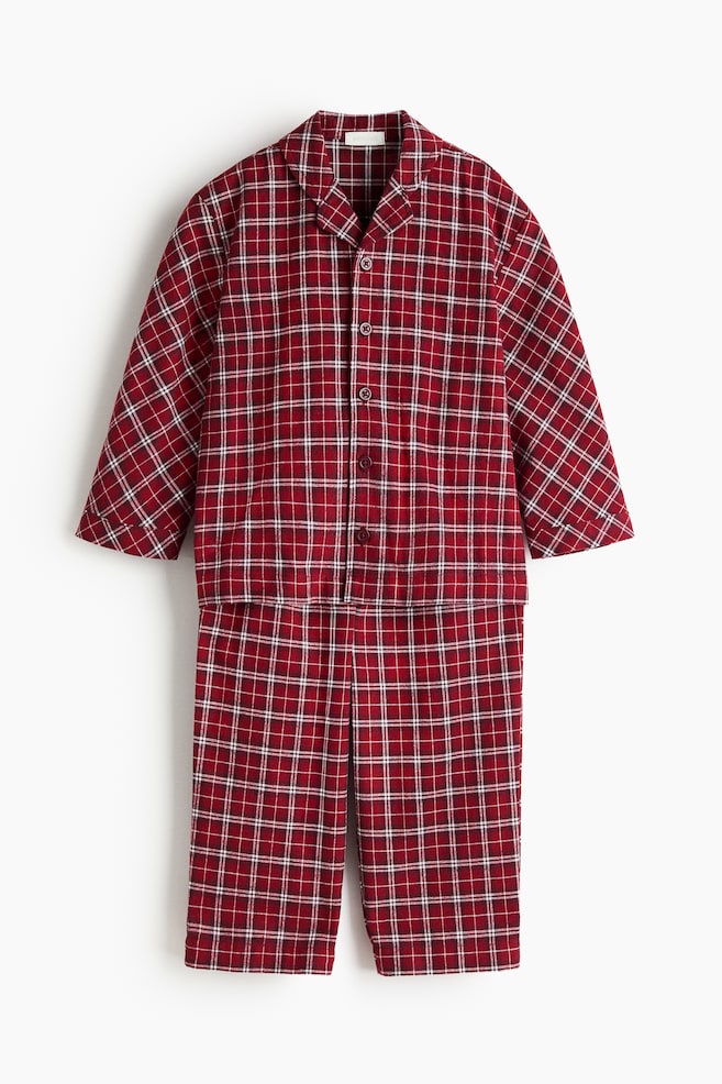 A two-piece pyjama set is shown from the front, featuring a matching long-sleeved top and straight-leg trousers in a red, white, and black plaid pattern. The button-down top features notch lapels and dark buttons, while the matching trousers hang below to complete the set. - 1
