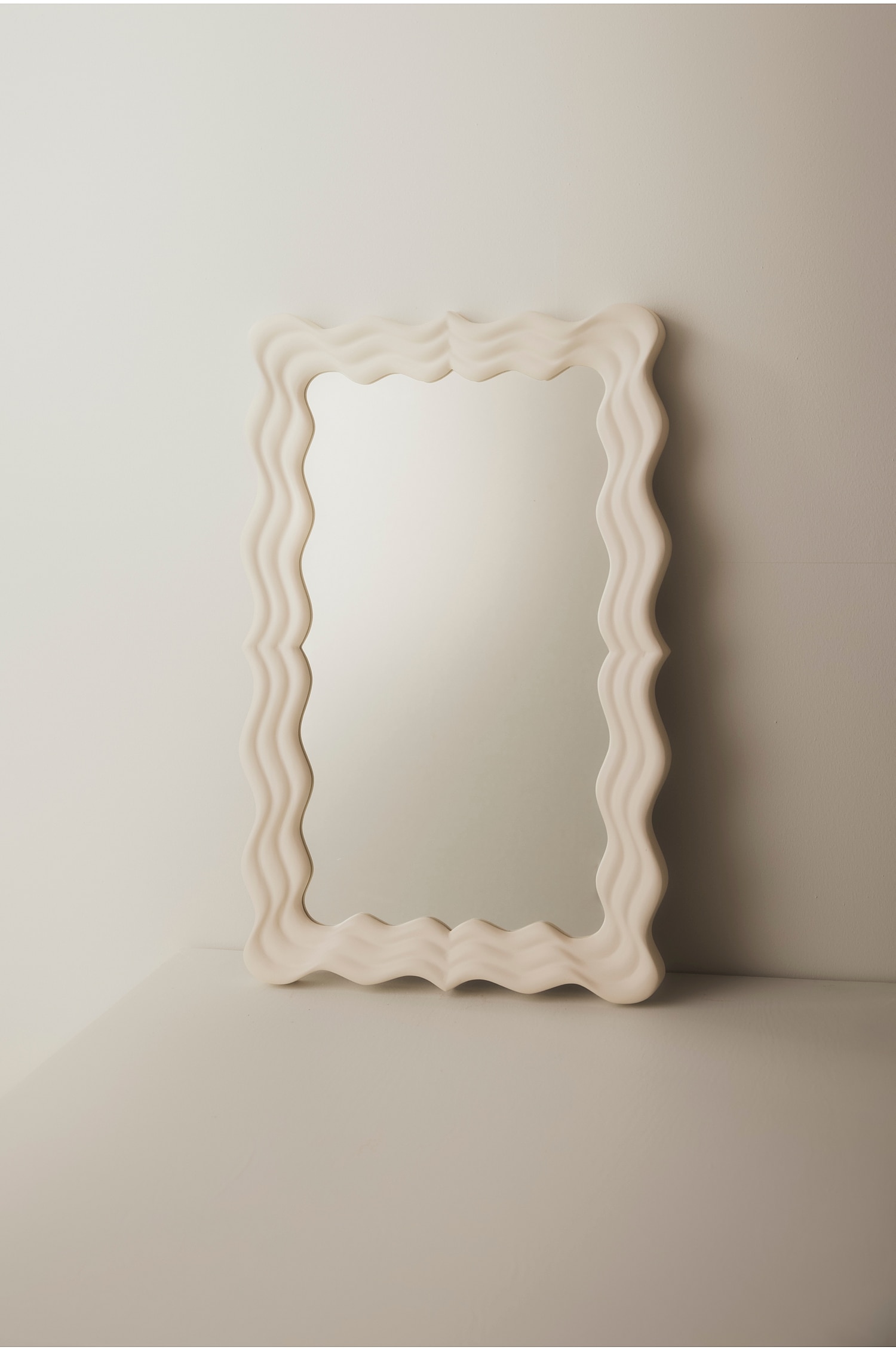 Een rechthoekige spiegel met een dikke, golvende lijst in een matte crÃ¨mekleur staat rechtop en is licht naar rechts gekanteld. De lijst vertoont een gebeeldhouwd, golvend patroon met gelaagde ribbels rond het centrale reflecterende glas.
