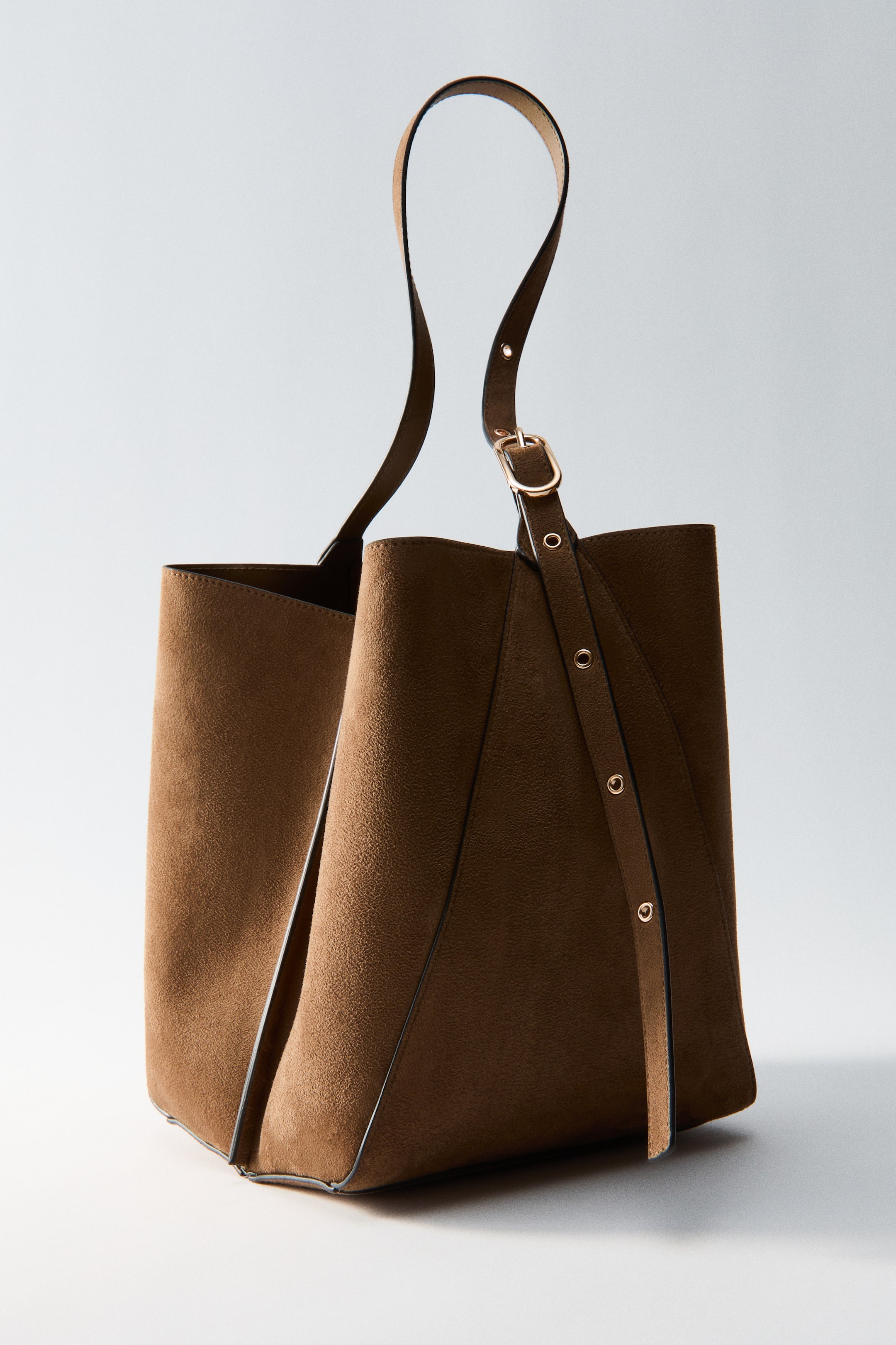 Voir une image plus grande: Un sac à bandoulière brun présente sa vue de face, mettant en valeur une texture semblable au suède et un haut ouvert. Une bandoulière réglable avec une boucle dorée et plusieurs œillets métalliques s'étend depuis le sac.