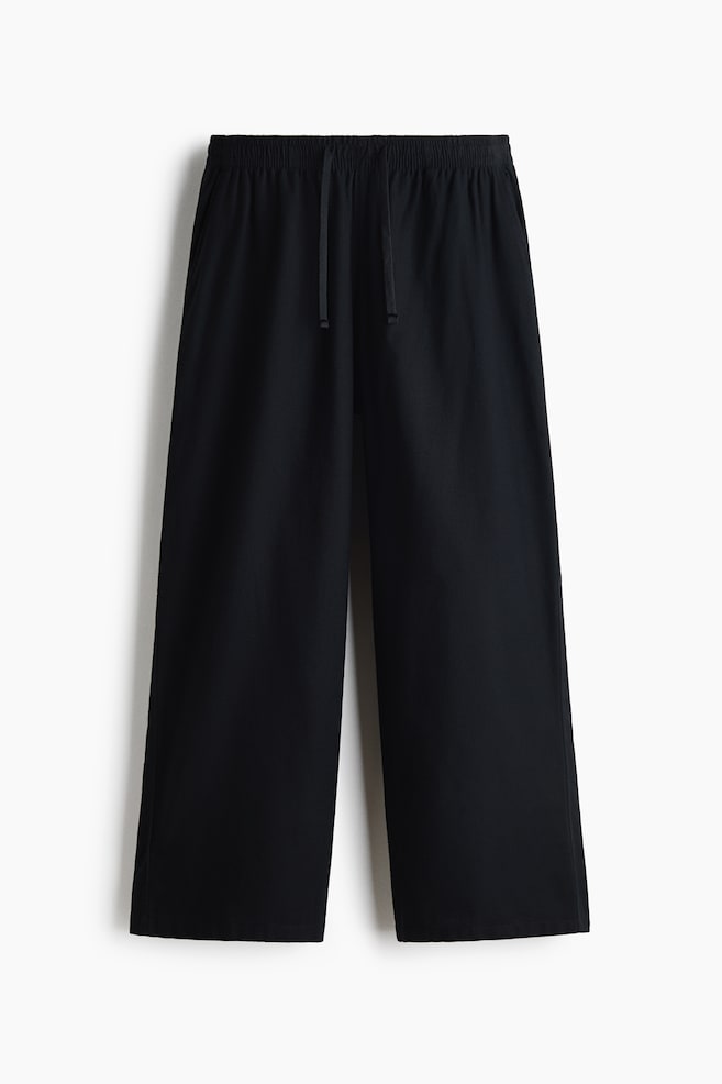 Un pantalon noir à jambe large est présenté de face, mettant en valeur une taille élastiquée avec des cordons de serrage assortis pendants au centre. Le pantalon affiche une coupe décontractée et ample qui se prolonge en jambes larges avec des ourlets droits. - 1