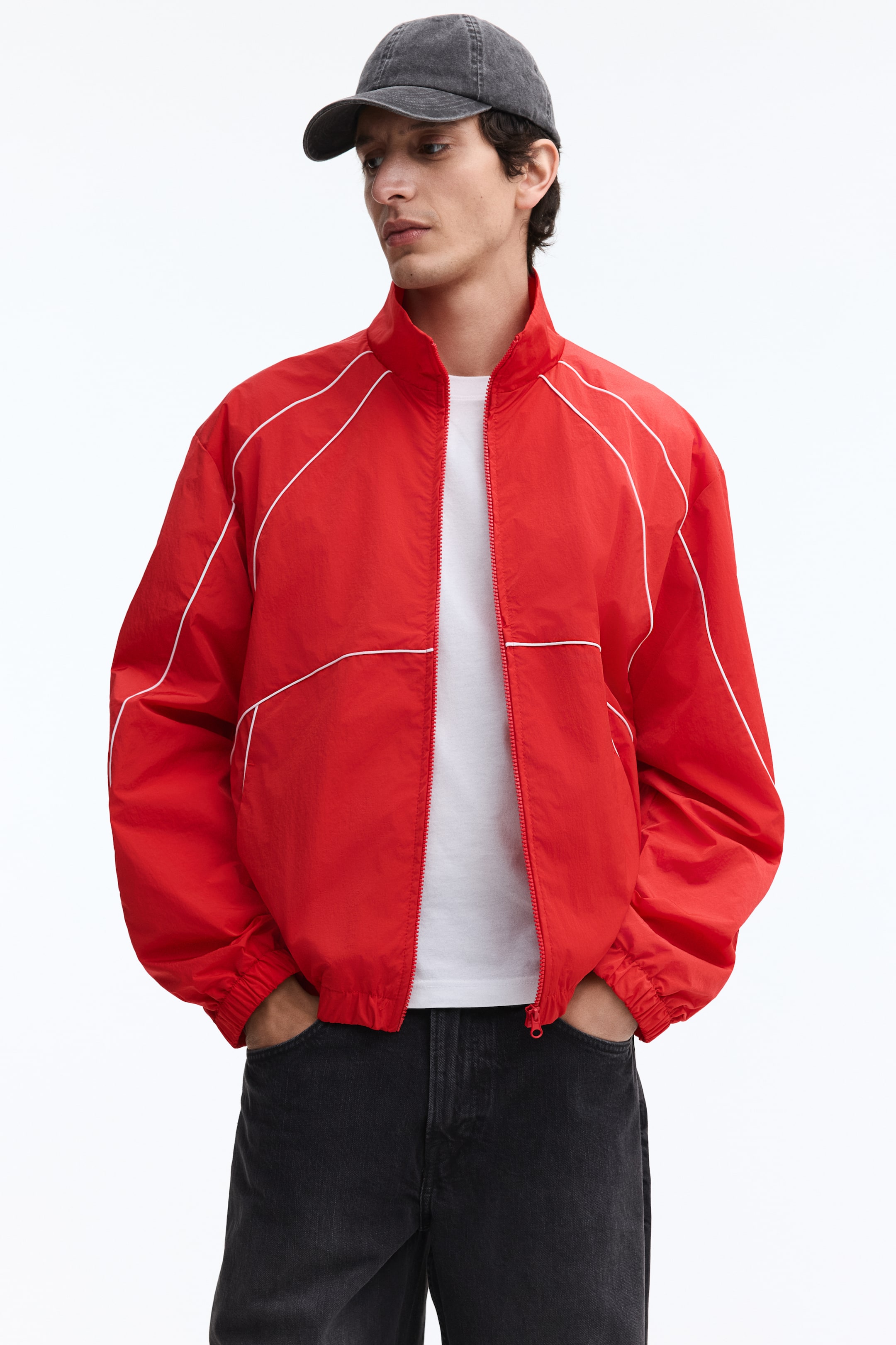 View larger image: The model is wearing a red, crisp nylon track jacket with white contrasting piping. It has a stand collar, a partially unzipped front that reveals a white t-shirt, and covered elastic at the cuffs and hem, with the model's hands resting in the side pockets.