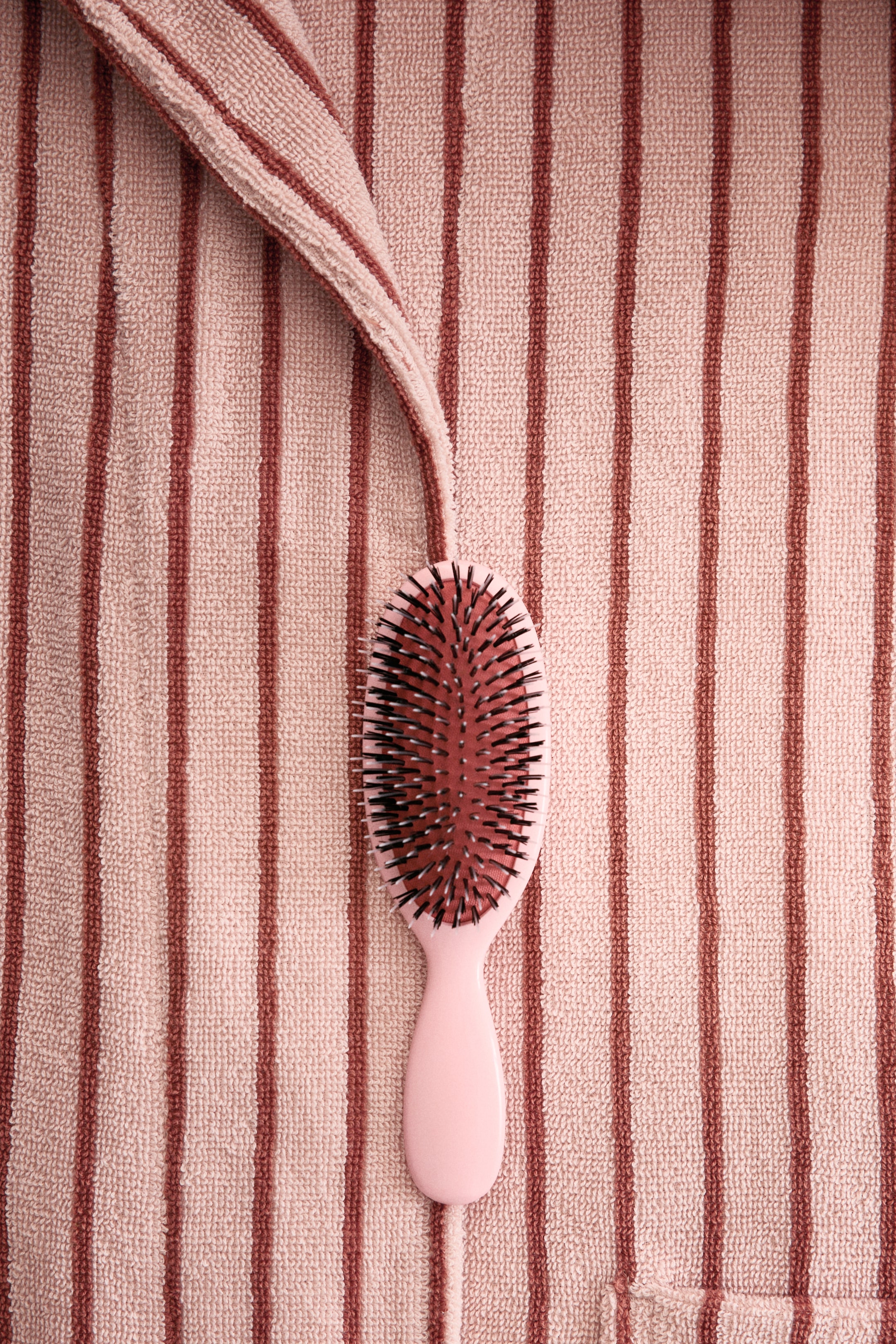 Ingrandisci l'immagine: Un accappatoio a righe mette in mostra il suo tessuto in spugna di cotone, caratterizzato da strisce verticali alternanti di rosa chiaro e scuro. Un bordo piegato appare in alto, mentre un angolo della tasca è visibile in basso a destra.