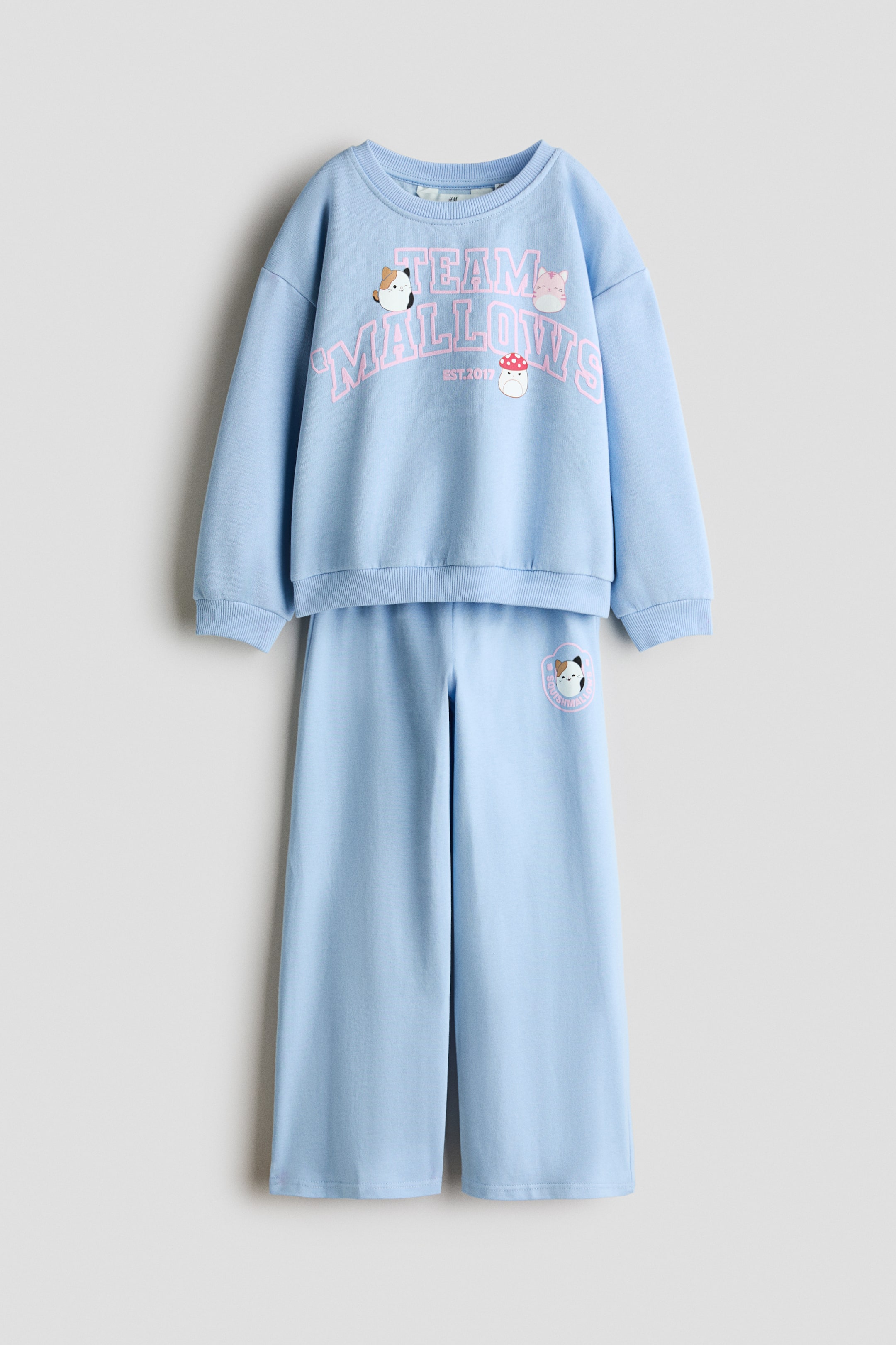 View larger image: A light blue two-piece lounge set is shown from the front, featuring a long-sleeved crew-neck sweater and matching flared leggings. The sweater features dropped shoulders, ribbed cuffs and hem, and a pink collegiate-style “TEAM MALLOWS” graphic on the chest, while the matching flared leggings display a small circular character print on the left thigh.