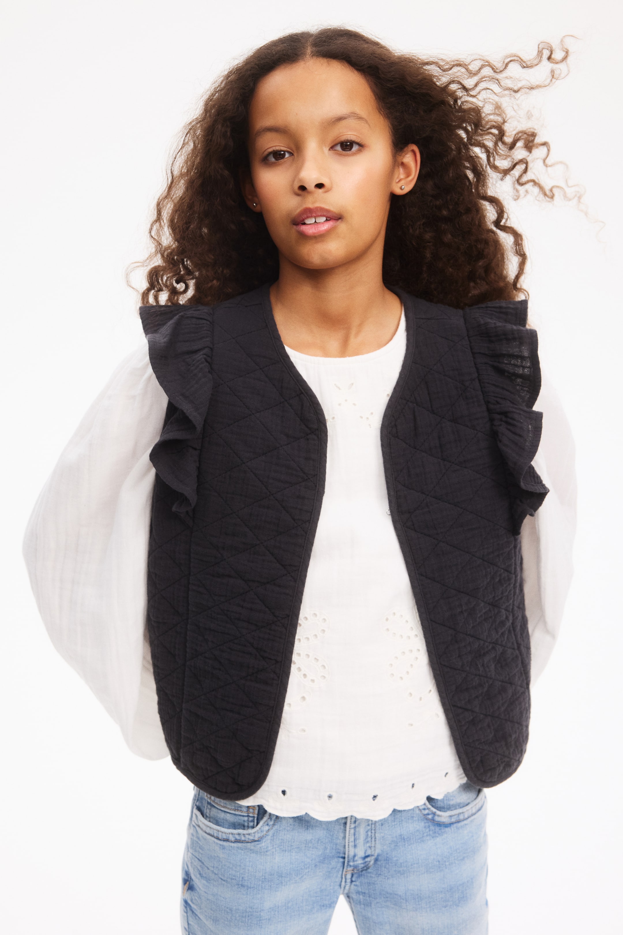 View larger image: A black quilted cotton muslin waistcoat features an open front with a V-shaped neckline, layered over a white top and finished with a flounce on the shoulders.