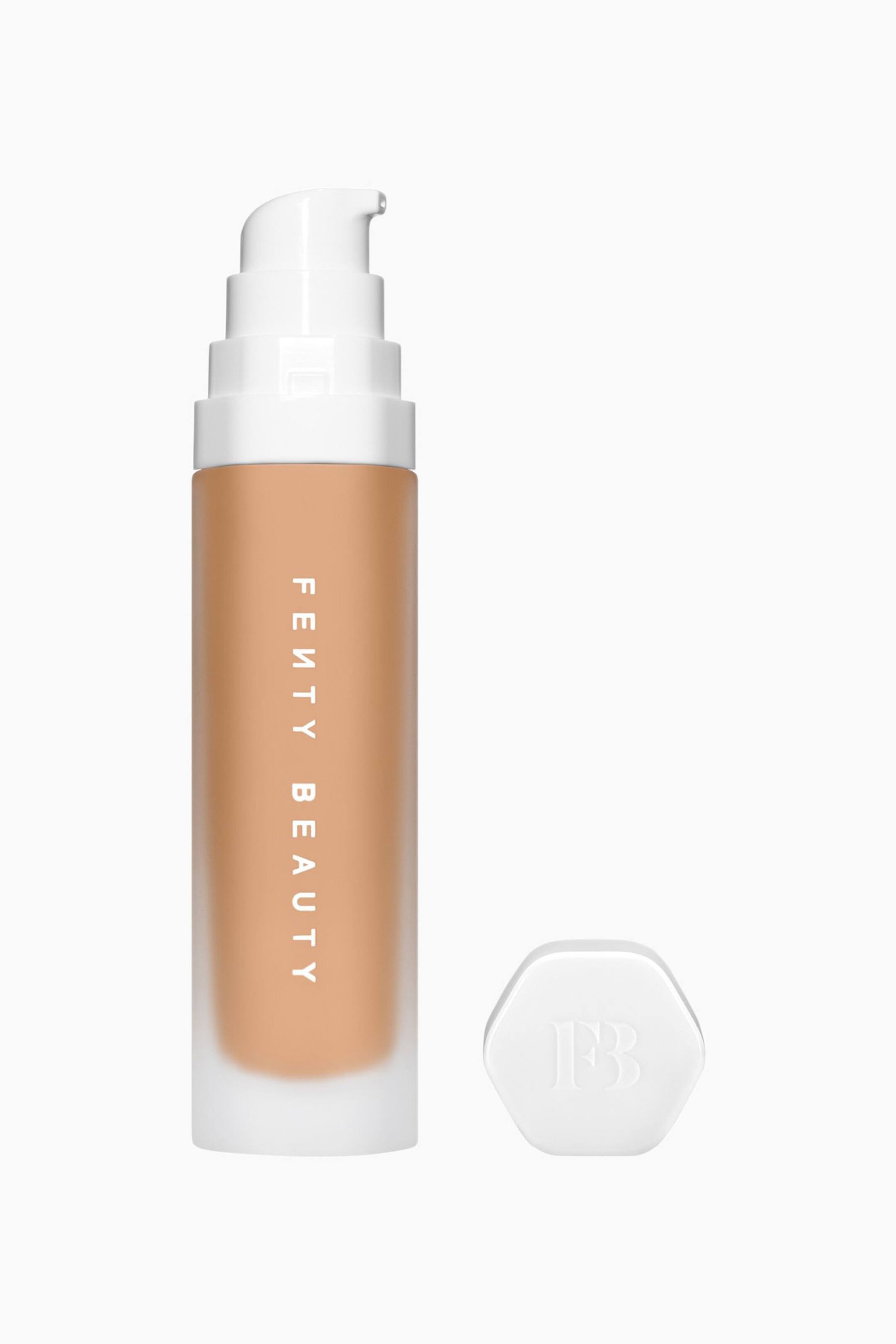 Visa större bild: Soft'lit Naturally Luminous Longwear Foundation står upprätt och visar upp en cylinderformad flaska i frostat glas fylld med en mediumtanfärgad flytande formula, med vertikal vit varumärkesmärkning. En vit pumpdispenser sitter på ovansidan, och till höger ligger en separat vit hexagonal kork med präglad "FB"-logotyp.