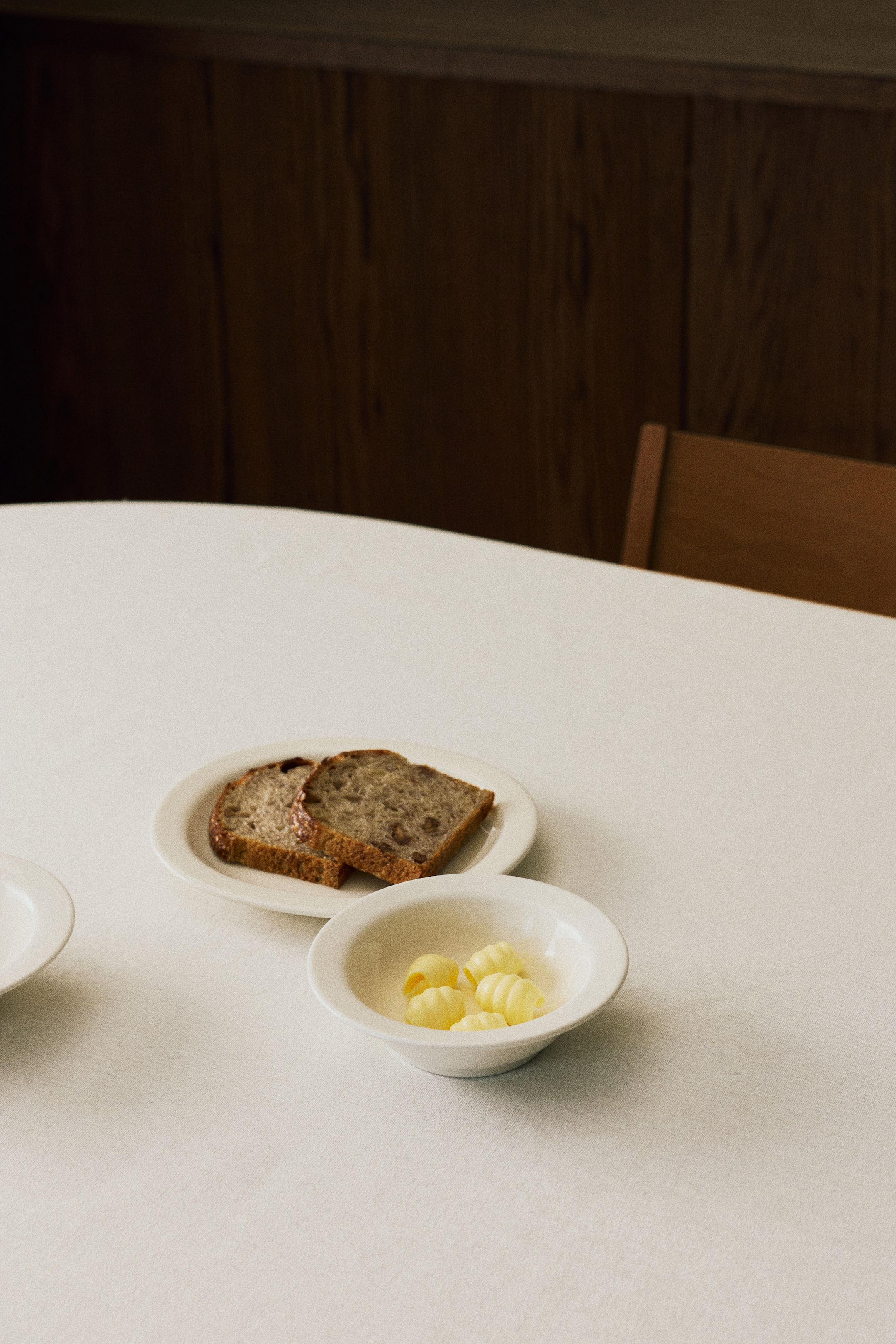 View larger image: A small porcelain bowl featuring a minimalist design with a classic wide rim is holding several yellow butter curls. A matching small porcelain plate is placed nearby, holding two slices of bread.
