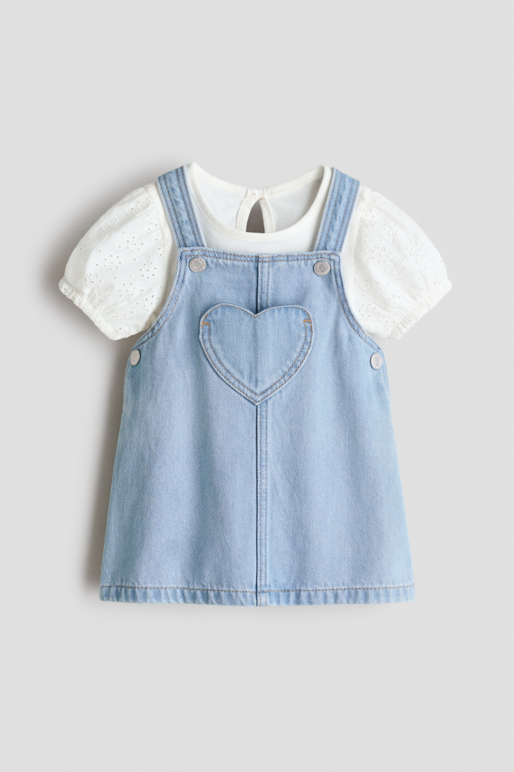 View larger image: A light blue dungaree dress is displaying its front, featuring a heart-shaped chest pocket, shoulder straps with press-stud fastenings, and press-studs at each side of the waist. A white top is layered underneath, presenting a round neckline with a keyhole opening at the back, and puff sleeves detailed with broderie anglaise and elasticated cuffs.