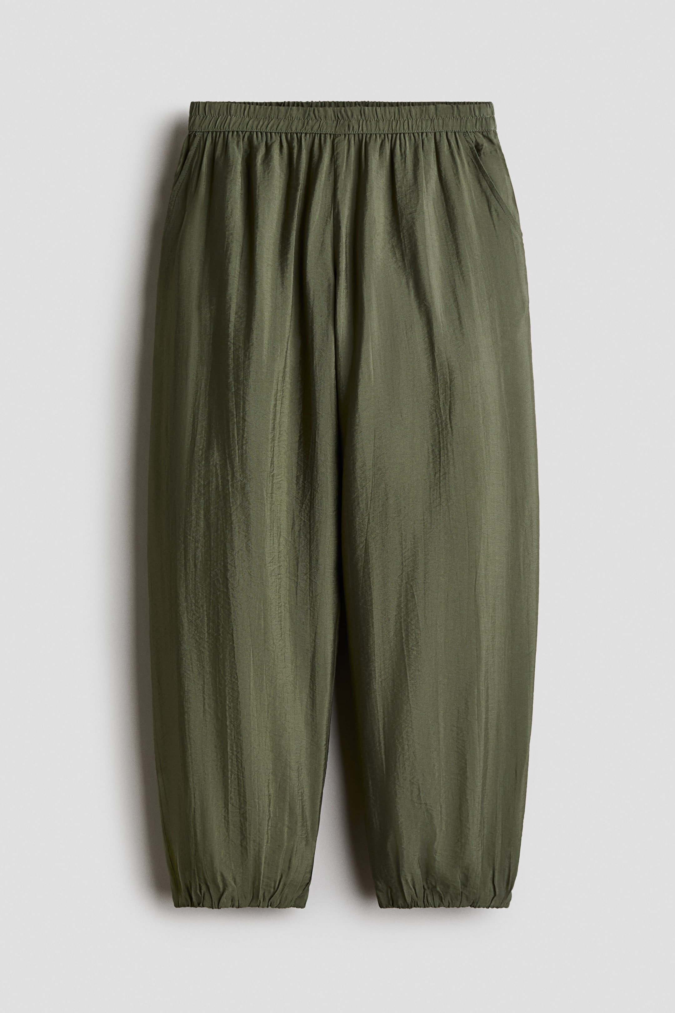 View larger image: Olive green trousers feature a gently crinkled viscose-blend weave with a subtle sheen. They are designed with an elasticated waist, diagonal side pockets, and balloon legs with elasticated hems.