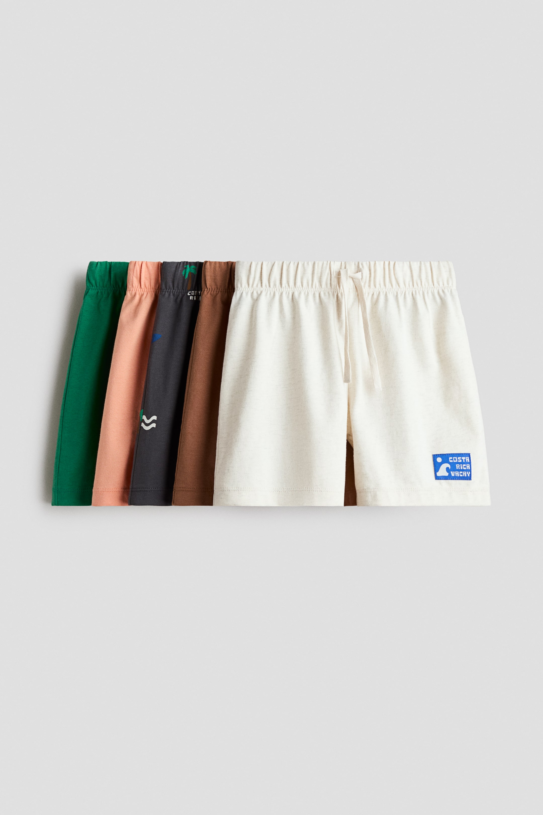 Agrandir l'image: Un ensemble de cinq shorts longueur genou est présenté dans une disposition en couches, avec des couleurs incluant le vert, le pêche, le gris foncé, le brun et le blanc cassé. Chaque paire est confectionnée dans un tissu jersey de coton doux, avec une taille élastiquée recouverte et un cordon de serrage pour un enfilage facile. Le short blanc cassé, placé à l'avant, arbore également un écusson graphique bleu sur le bas de la jambe droite.