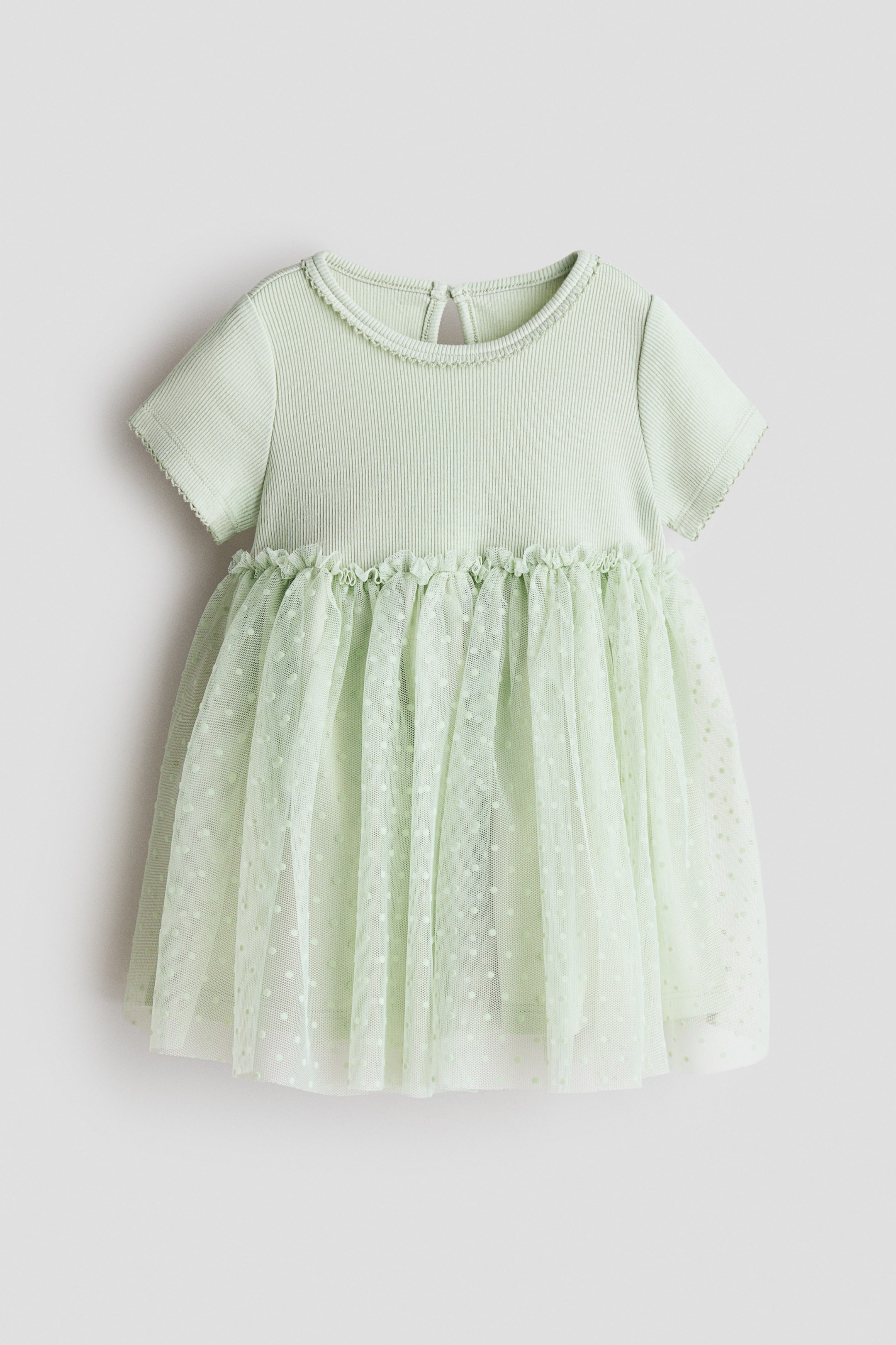 Ampliar la imagen: Vestido verde claro de manga corta que muestra su parte frontal, con corpiño de jersey acanalado, escote redondo rematado con picot y mangas cortas también rematadas con picot; frunces con volantes definen la cintura y dan paso a una falda de tul forrada y acampanada con estampado de puntos en foil, y en la parte posterior del escote se aprecia una abertura en forma de ojo de cerradura con botón.
