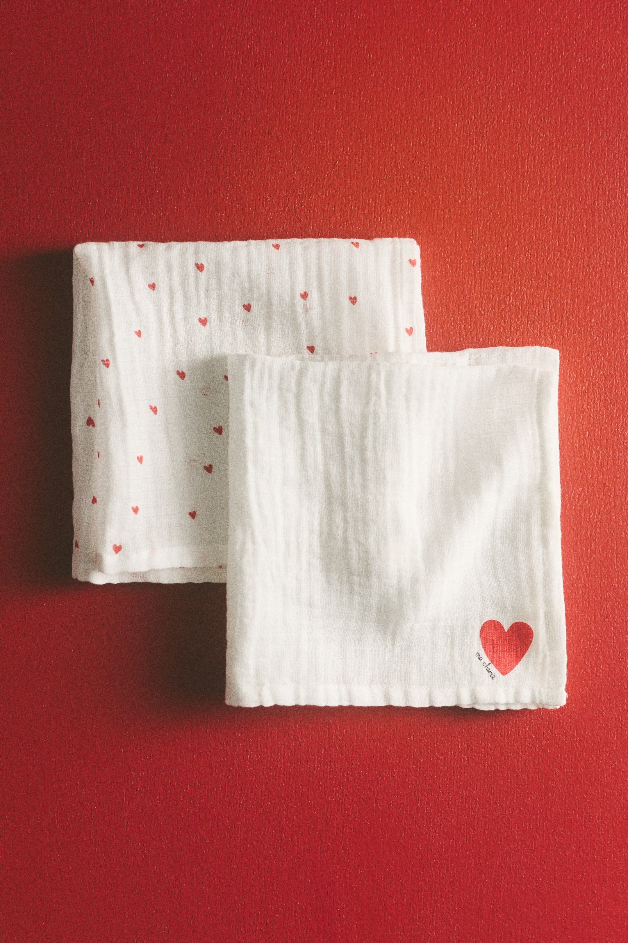 View larger image: Two white, textured fabric cloths are laid flat and overlapping. One cloth is adorned with small red hearts, while the other is plain white, showcasing a large red heart with "ma chérie" text in its bottom right corner.