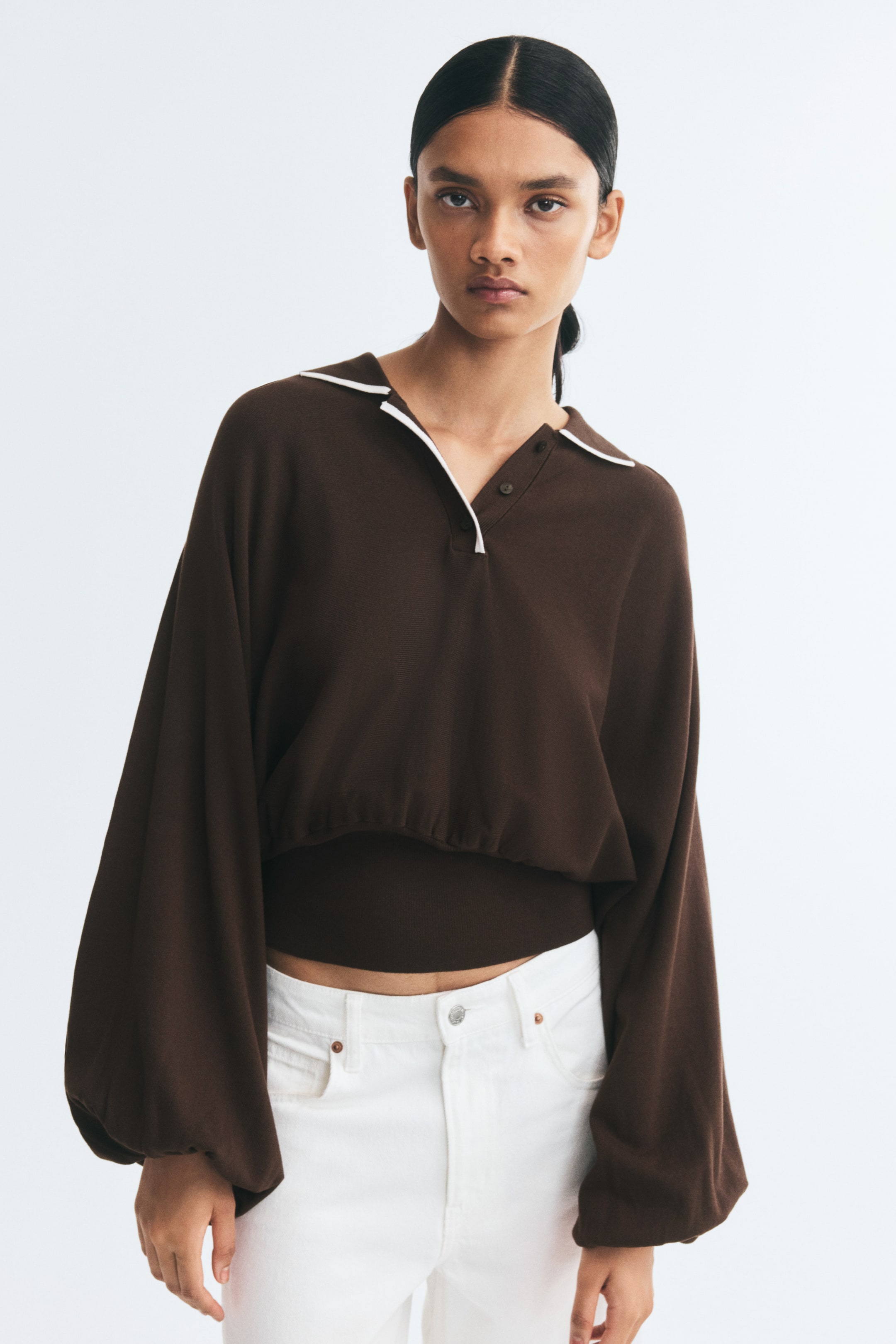View larger image: The model is wearing a brown knit polo sweater, featuring a white-trimmed collar, button placket, and long, voluminous batwing sleeves. The sweater has a wide, fitted waistband with gathers along the top edge and is paired with white jeans.