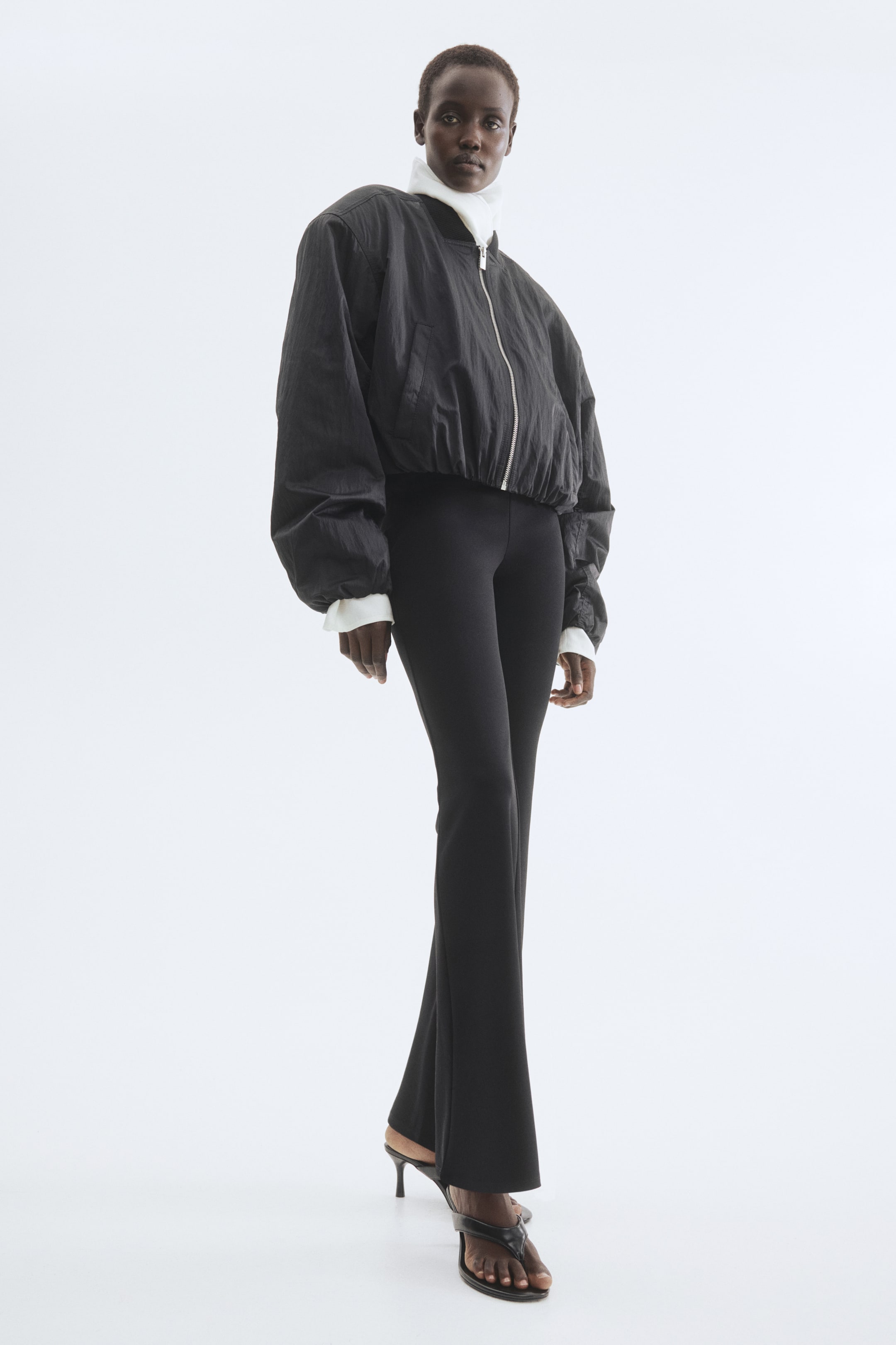 Ingrandisci l'immagine: Una giacca bomber nera corta presenta una texture stropicciata, maniche voluminose e una zip frontale argentata, stratificata su un dolcevita bianco con i polsini bianchi che si estendono dai polsini elasticizzati della giacca. Pantaloni neri a vita alta a zampa d'elefante aderiscono in modo aderente alla coscia e si allargano verso l'orlo, poggiando su sandali neri con tacco.