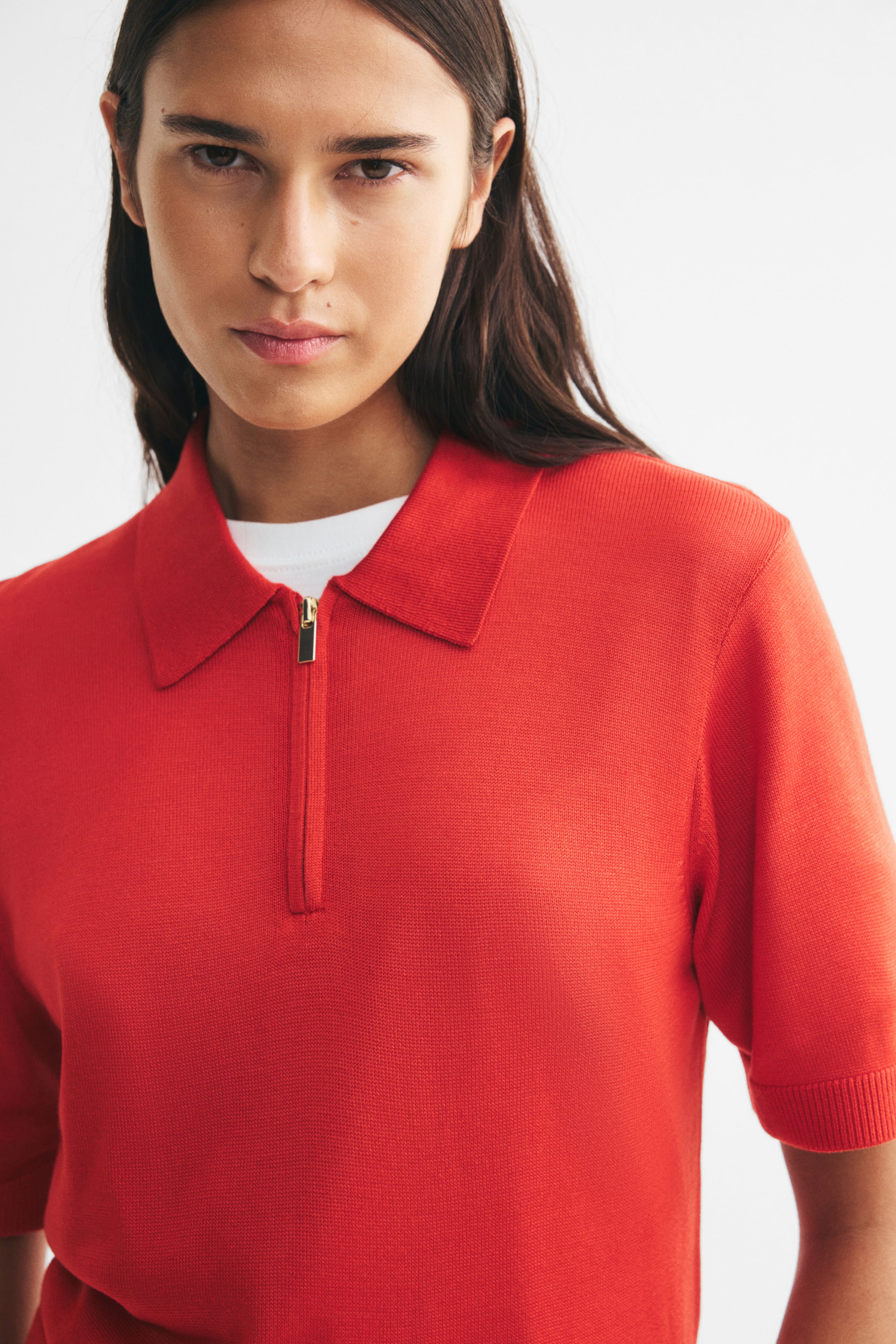 View larger image: The model is wearing a red fine-knit polo shirt with a collar and a gold-toned half-zip. The short-sleeve shirt features ribbed cuffs and is layered over a white crew-neck T-shirt.