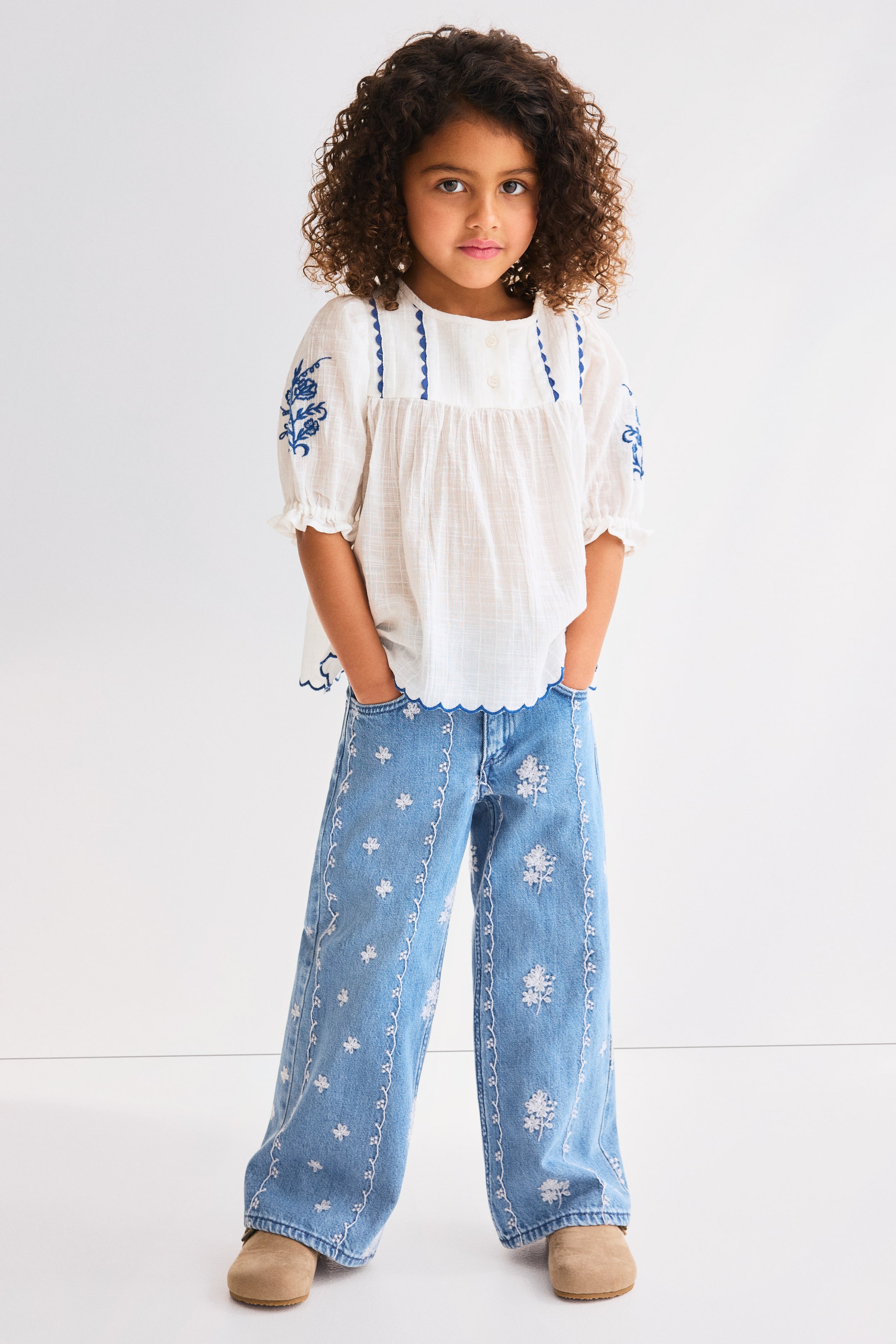 View larger image: A white textured blouse features a gathered bodice, ruffled short sleeves, and blue floral embroidery on the puffed sleeves and yoke, complemented by a blue scalloped trim along the yoke and hem. It is paired with light blue wide-leg jeans, adorned with white embroidered floral and vine patterns along both legs and a scalloped waistband.