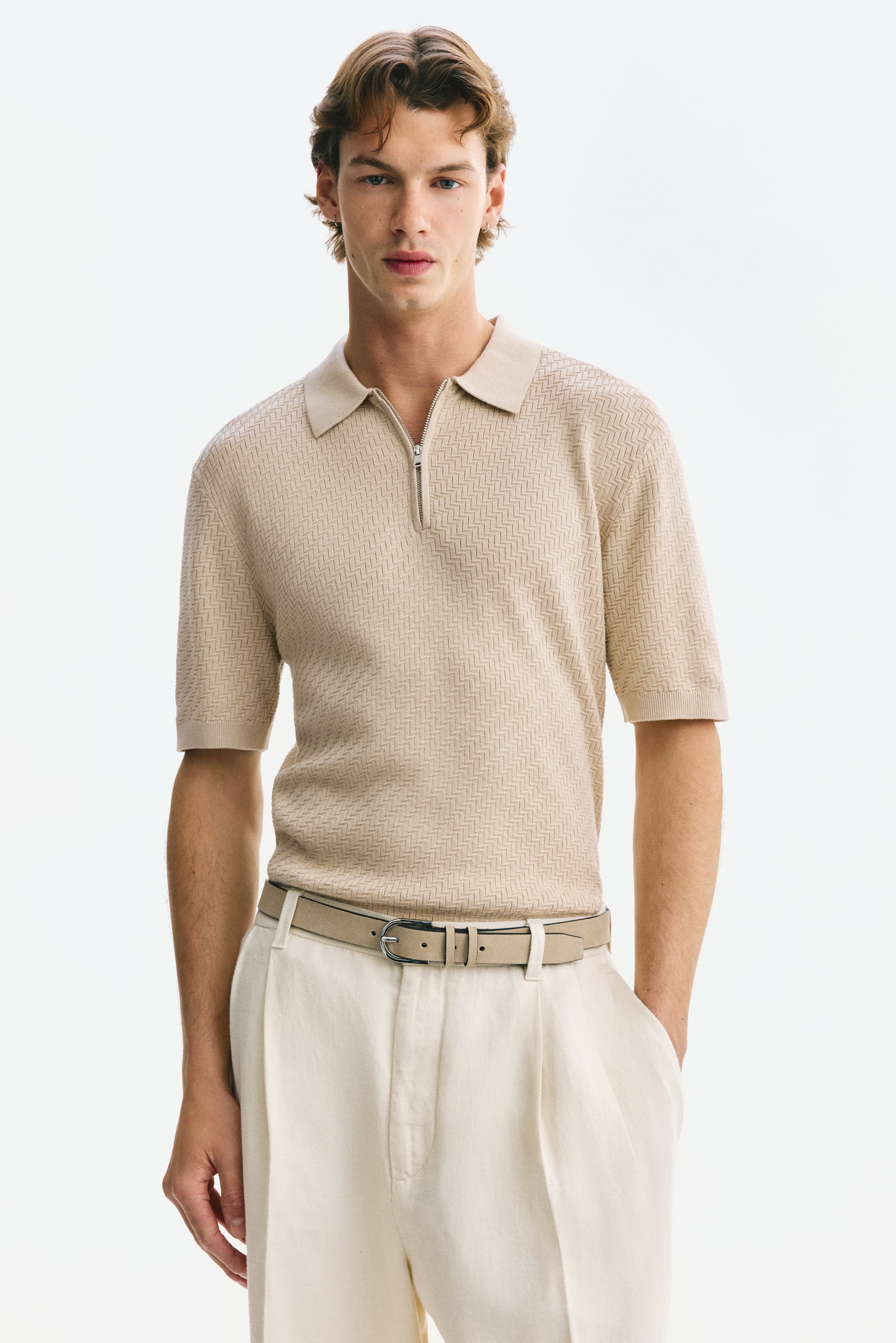 Ver imagen más grande: Una camiseta polo de manga corta en color beige, que presenta un patrón de tejido texturizado, un cuello acanalado, un cierre de media cremallera en el escote, puños acanalados y un dobladillo acanalado, es llevada por el modelo, ajustándose ceñidamente al cuerpo y metida en unos pantalones claros.
