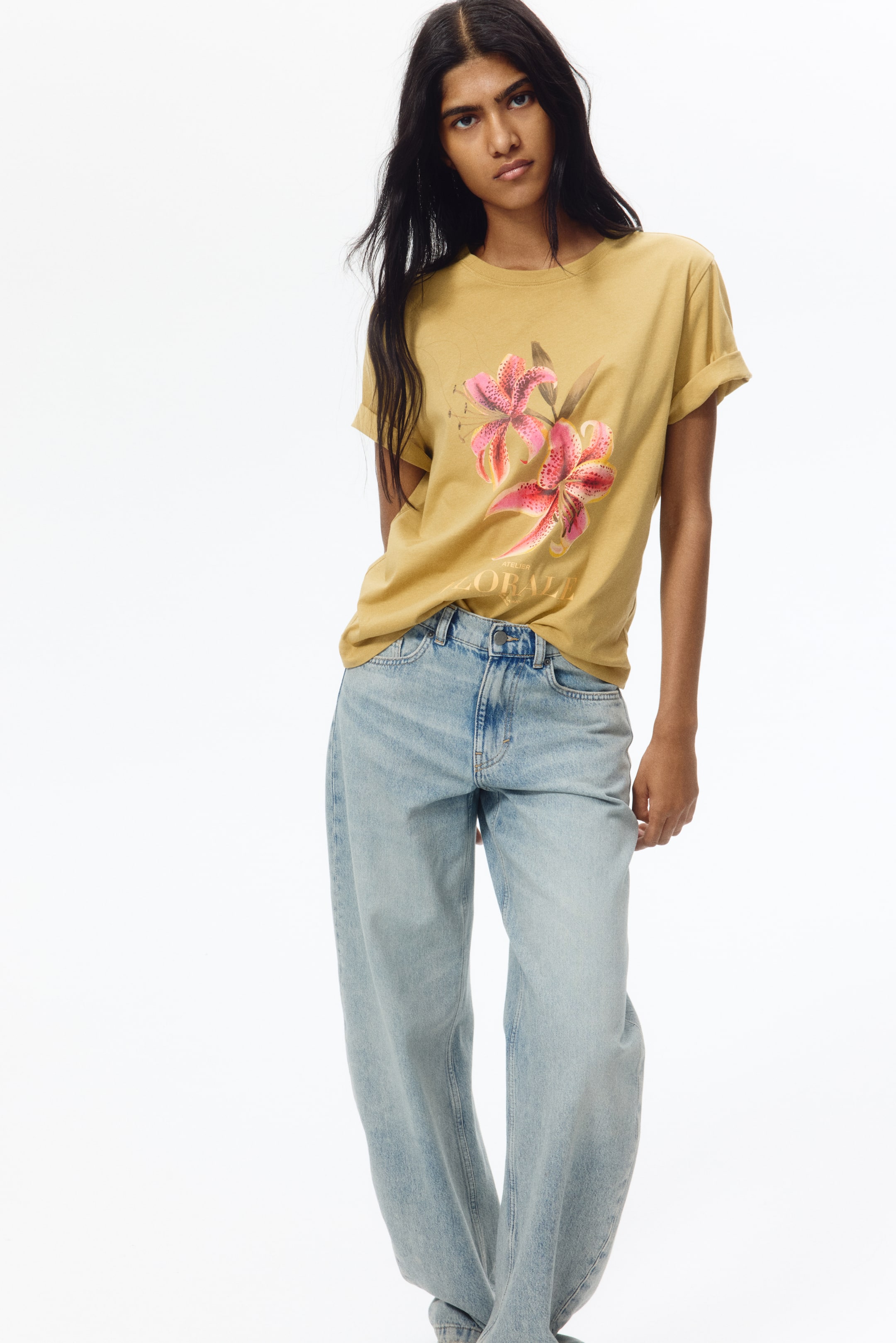 View larger image: A mustard yellow t-shirt showcases a graphic print of pink and red lilies with the partially visible text 'ATELIER FLORALE' on the front. The short sleeves are rolled up, and the hem is partially tucked in, offering a relaxed fit.