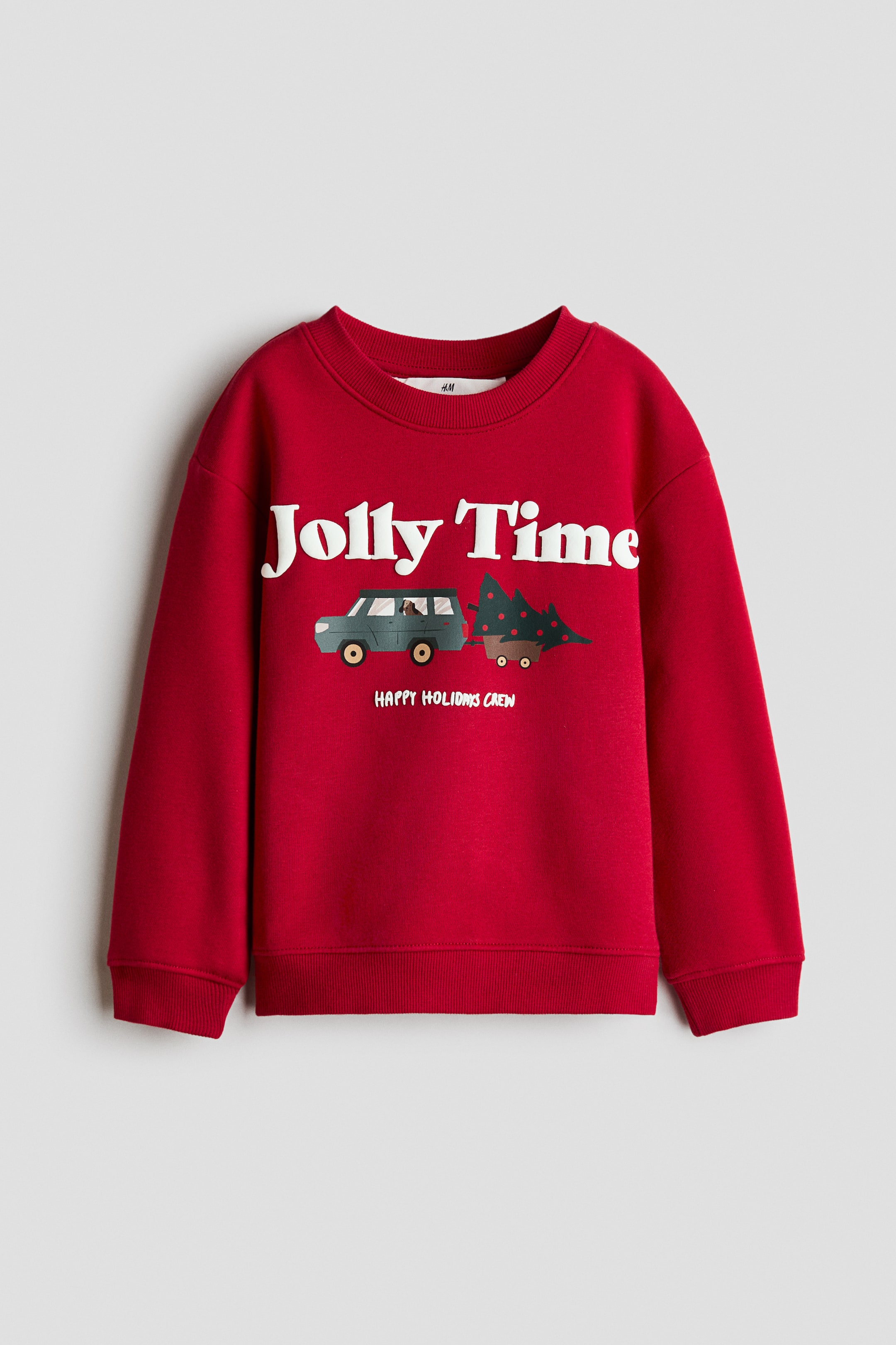 View larger image: A red sweatshirt is shown from the front, featuring a ribbed crew neckline, dropped shoulders, long sleeves, and ribbed hem and cuffs. The front displays a printed graphic with the white text "Jolly Time" above an illustration of a utility vehicle towing a decorated evergreen tree, with the smaller text "HAPPY HOLIDAYS CREW" printed underneath.