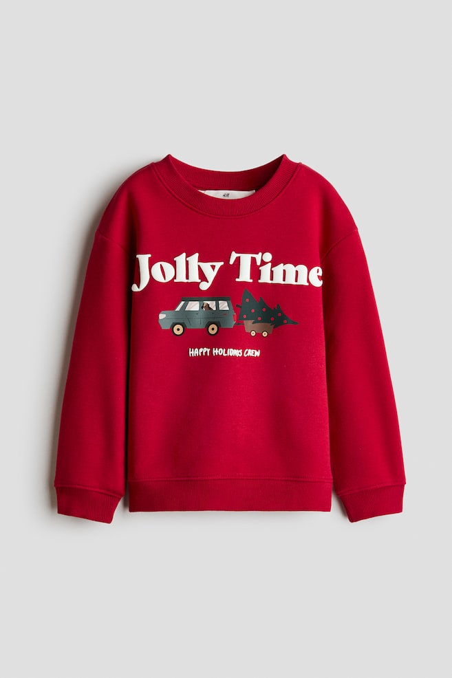 A red sweatshirt is shown from the front, featuring a ribbed round neckline, dropped shoulders, long sleeves, and a ribbed hem and cuffs. The front displays a printed graphic with the white text "Jolly Time" above an illustration of a utility vehicle towing a decorated evergreen tree, with the smaller text "HAPPY HOLIDAYS CREW" printed underneath. - 1
