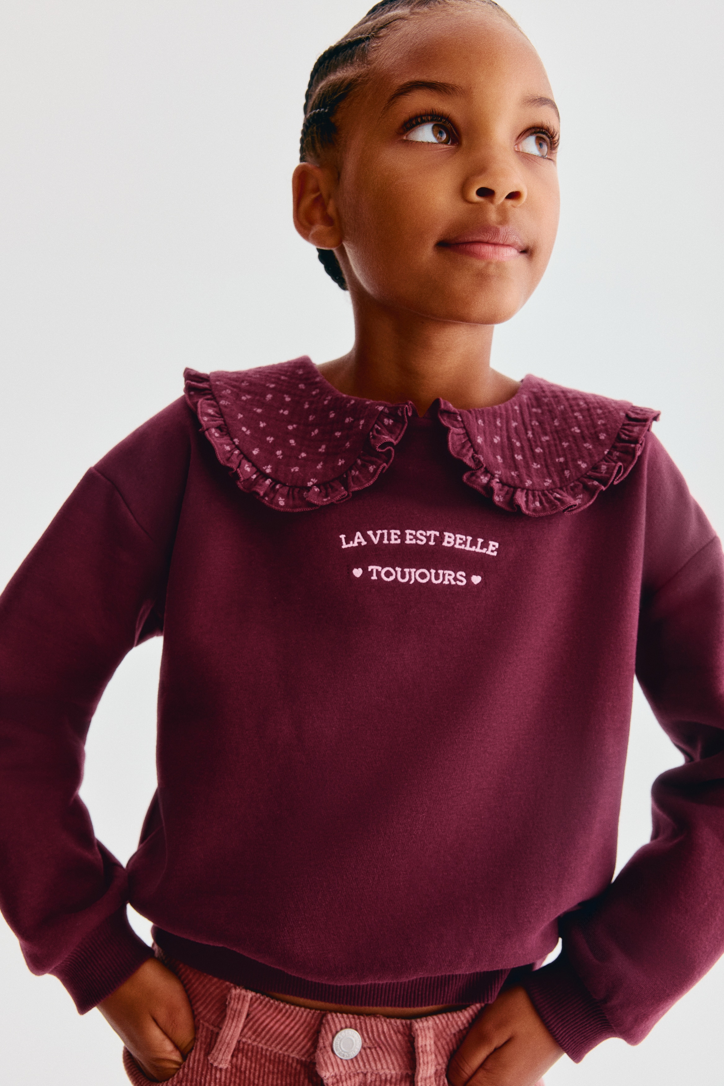 A front-facing burgundy sweater in brushed fleece features a ruffled, patterned Peter Pan collar and white embroidered text on the chest. The design showcases dropped shoulders, long sleeves with ribbed cuffs, and a ribbed hem.