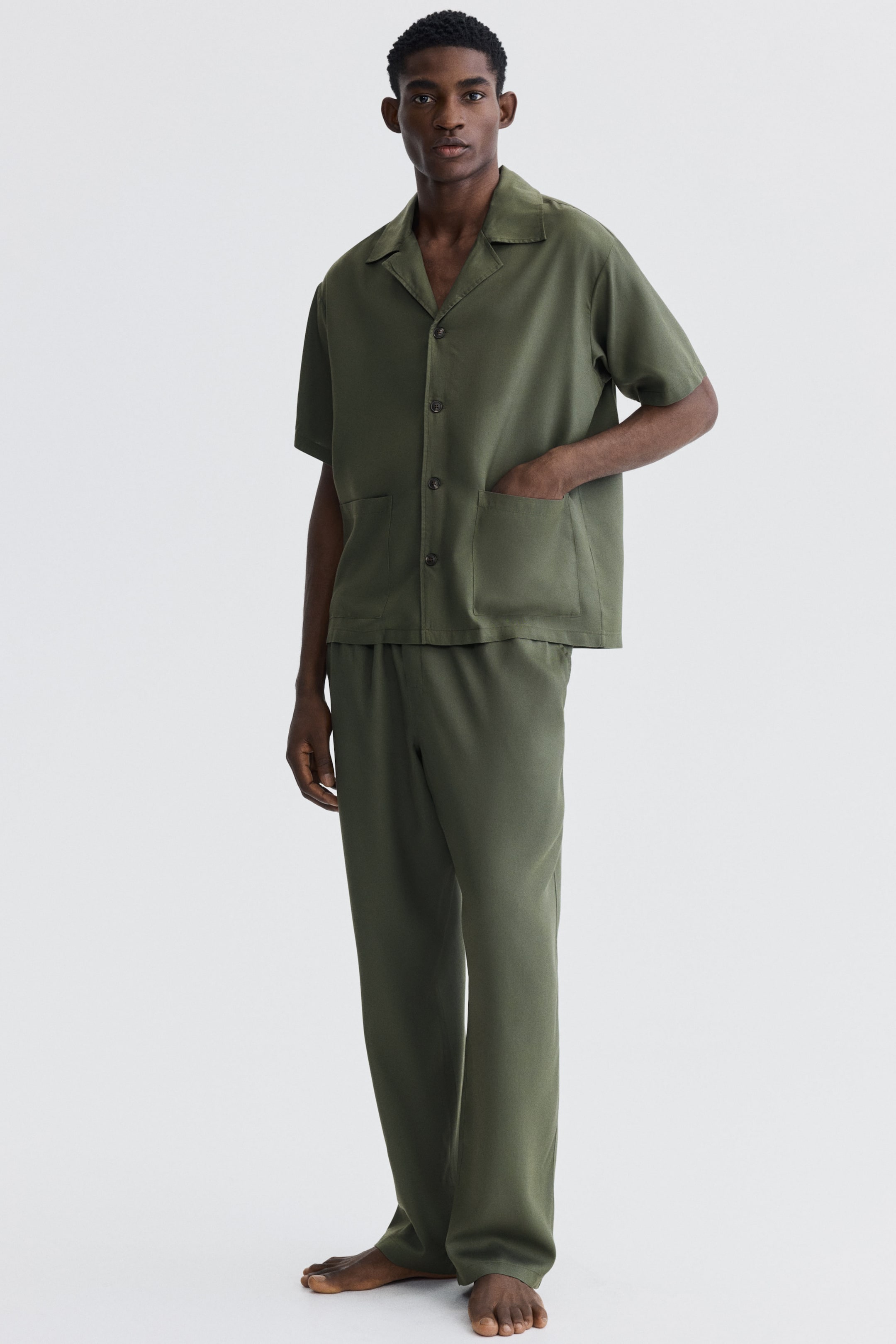 Ver imagen más grande: La modelo lleva una camisa de manga corta en color verde oliva, confeccionada en un tejido suave de lyocell. La prenda presenta un cuello resort, cierre frontal francés, dos bolsillos de parche en la parte delantera y un dobladillo recto con un ajuste relajado. Una de las manos de la modelo descansa en un bolsillo delantero.