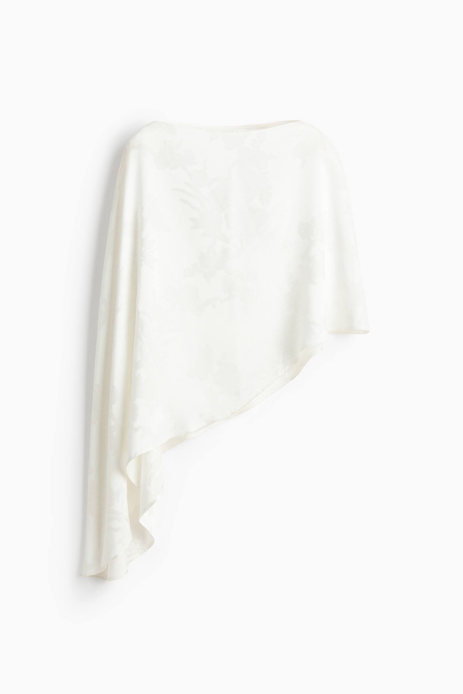 A cream-coloured cape in a jacquard weave with a subtle floral pattern and a slight sheen is hanging, featuring a boat neckline and a dramatic asymmetric hem that drapes longer on one side.