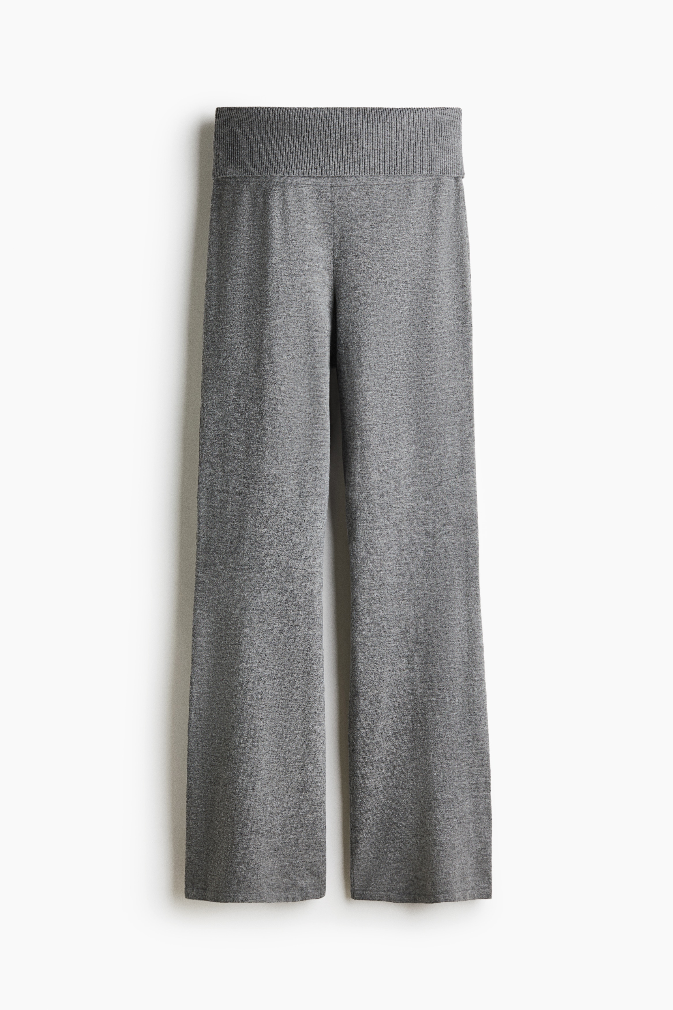 Eng anliegende Hose aus weichem grauem Strick mit umgeschlagenem Bund und ausgestellten Hosenbeinen, von dem Model getragen. Das Model hebt den Saum eines passenden gerippten Strickpullovers an, um den breiten Bund der Hose zu zeigen.