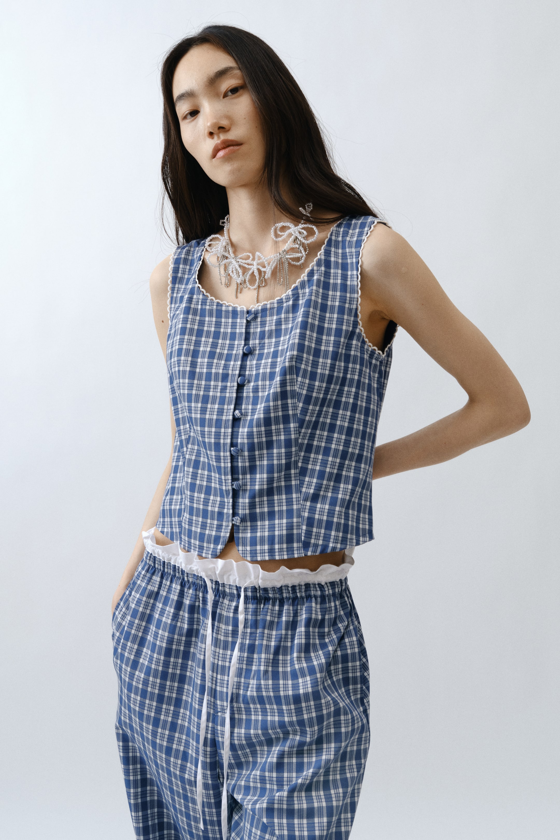 View larger image: A blue and white plaid sleeveless top at matching trousers ay ipinapakita mula sa bahagyang angled na front view. Ang top ay may round neckline na may scalloped white trim sa gilid at may blue front buttons, habang ang cropped hem nito ay nagpapakita ng white ruffled trim sa ilalim. Ang trousers ay may elasticated waistband na may nakikitang white ruffled trim at may white drawstring.