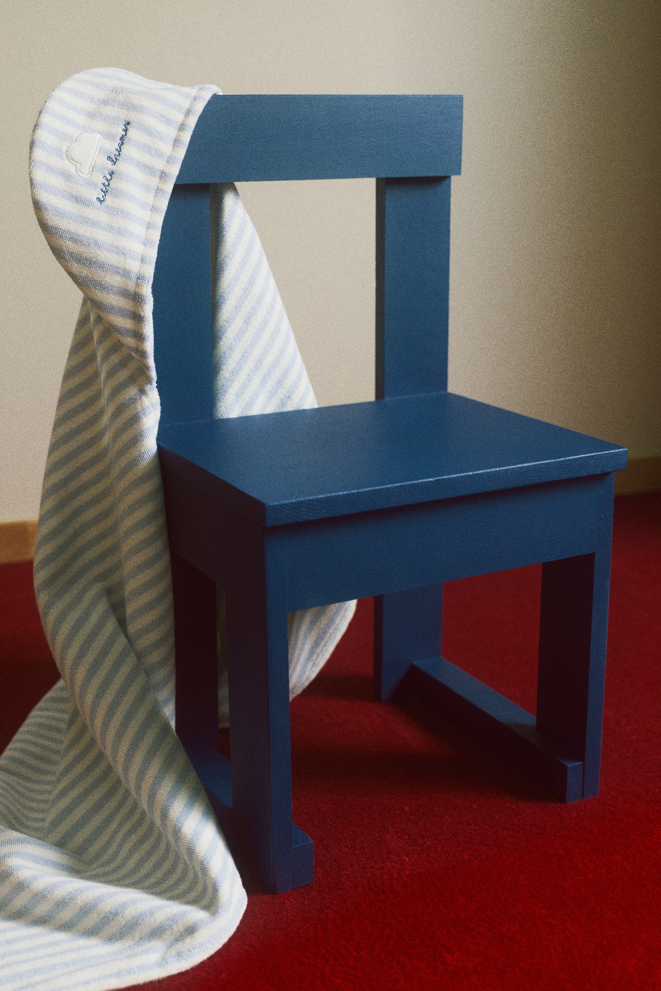 View larger image: A dark blue wooden chair is facing slightly right, featuring a square seat and a backrest with a single horizontal slat. A light blue and white striped blanket, embroidered with a cloud and "little dreamers," is draping over the chair's backrest.