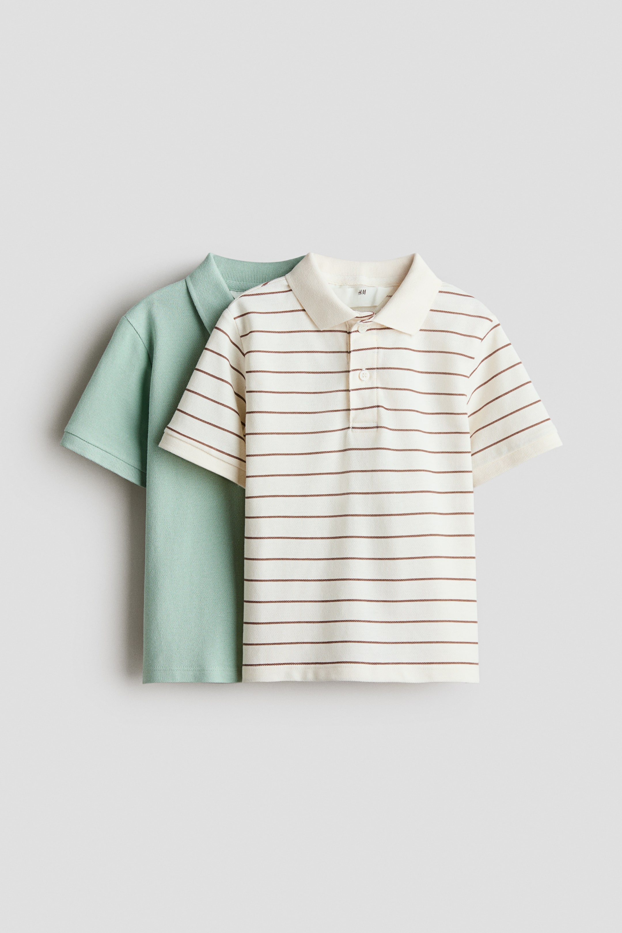 View larger image: Two polo shirts are positioned facing forward, with one slightly overlapping the other. The foremost shirt is cream with brown horizontal stripes, featuring a polo collar, a two-button placket, and short sleeves with ribbed cuffs. A solid light green polo shirt is partially visible in the background.