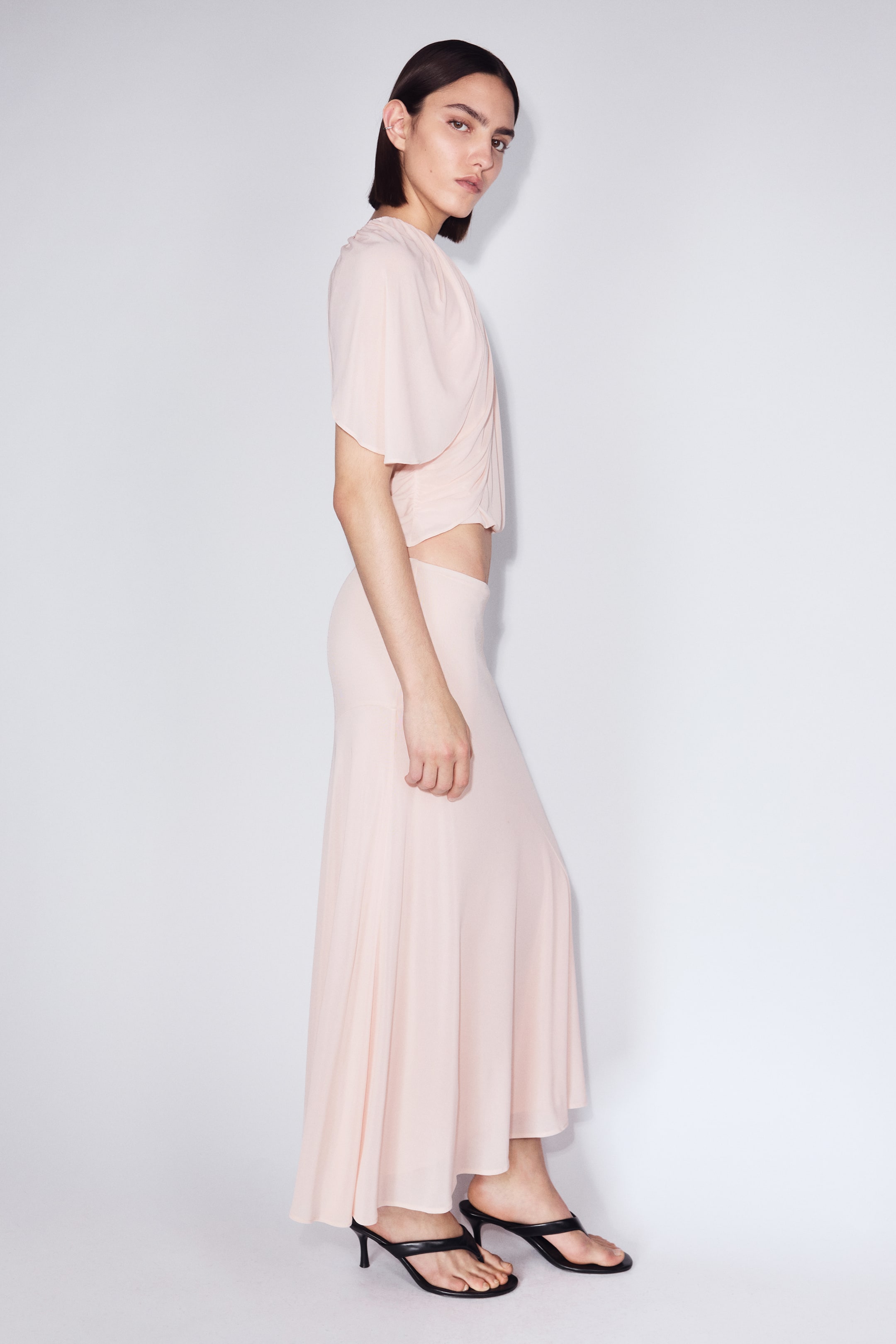 Agrandir l'image: Une tenue deux pièces rose pâle est présentée sous un angle latéral. Le haut court arbore des manches courtes drapées et un froncement latéral, tandis que la jupe longue assortie affiche une silhouette fluide et douce en ligne A.